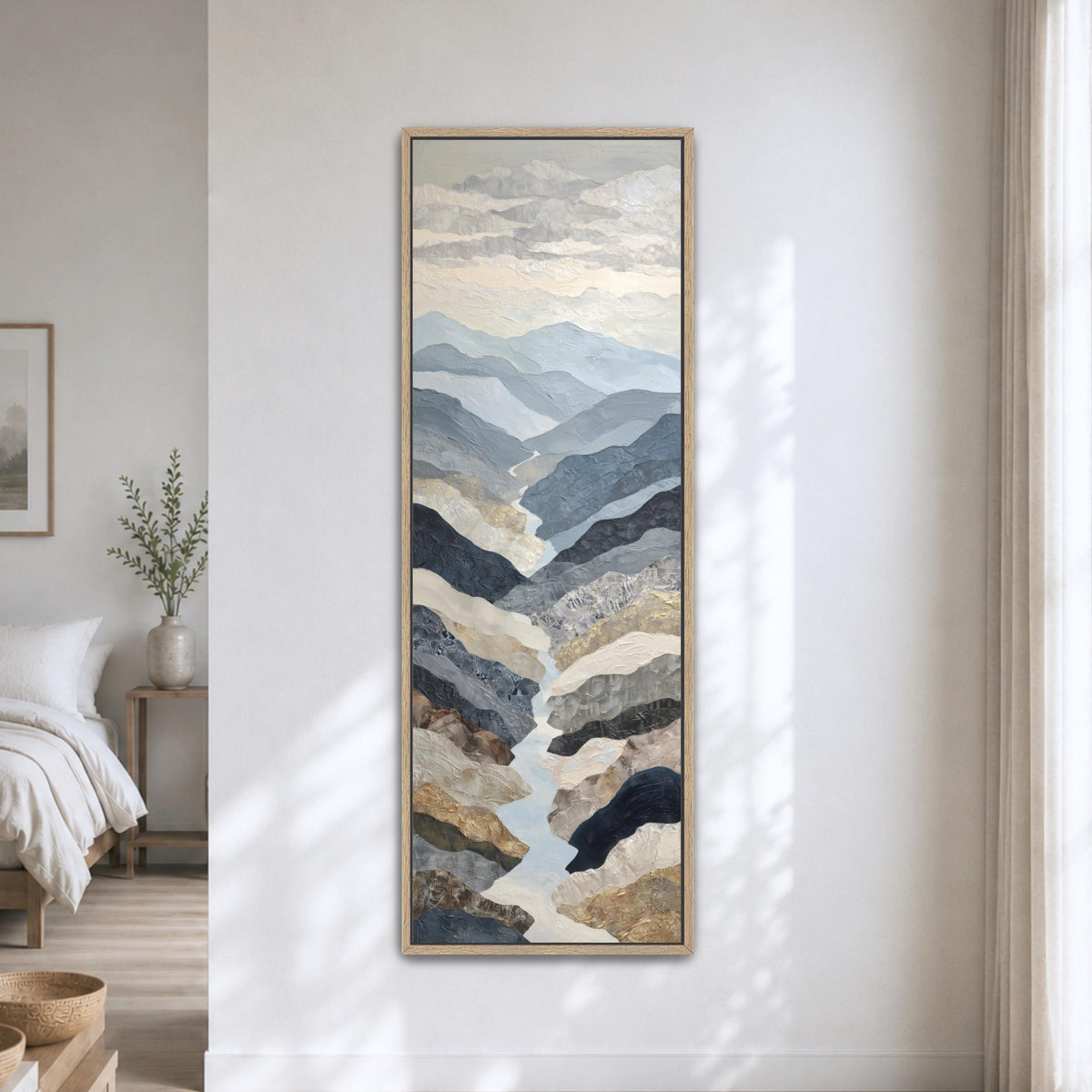 a large, framed painting of mountains with a winding river or stream running through it, hanging on a white wall in a room with a bed, a plant, and a window.