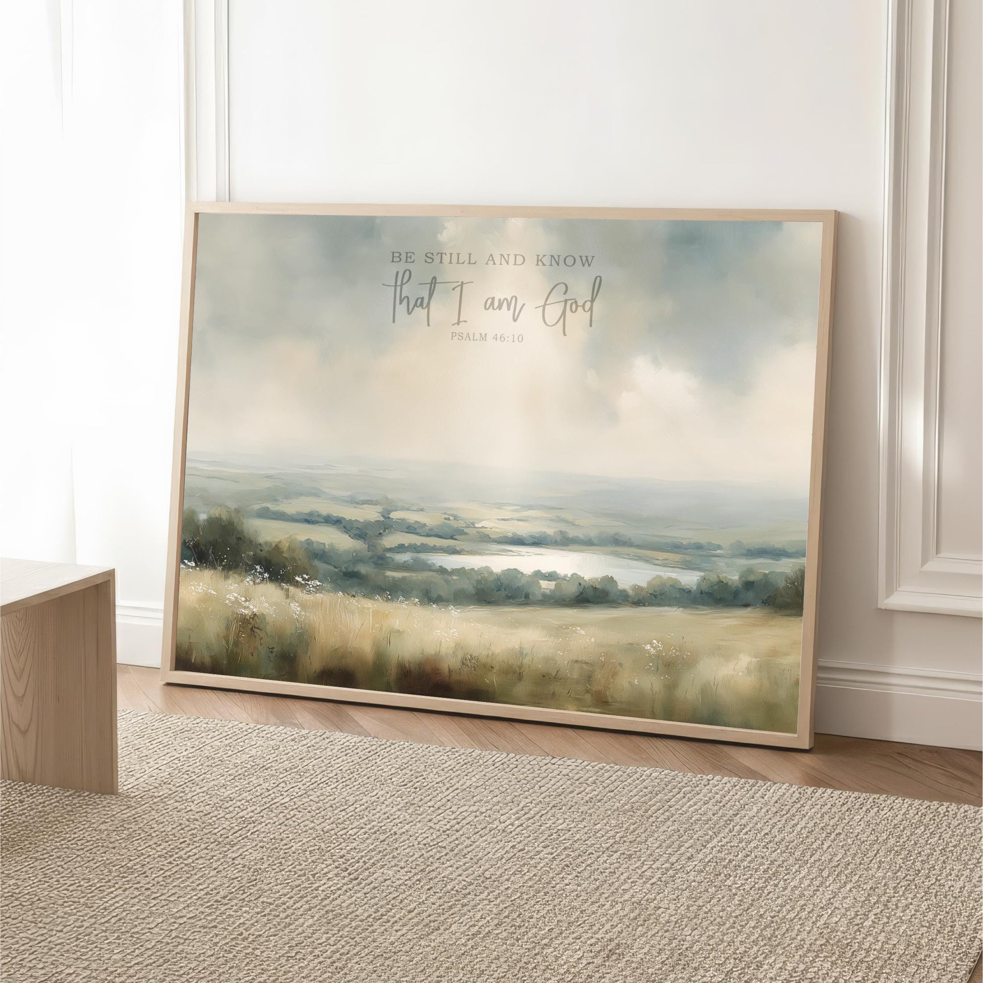 A framed landscape painting depicting a serene countryside scene with a lake in the distance, accompanied by the text "Be Still and Know That I am God" from Psalm 46:10.