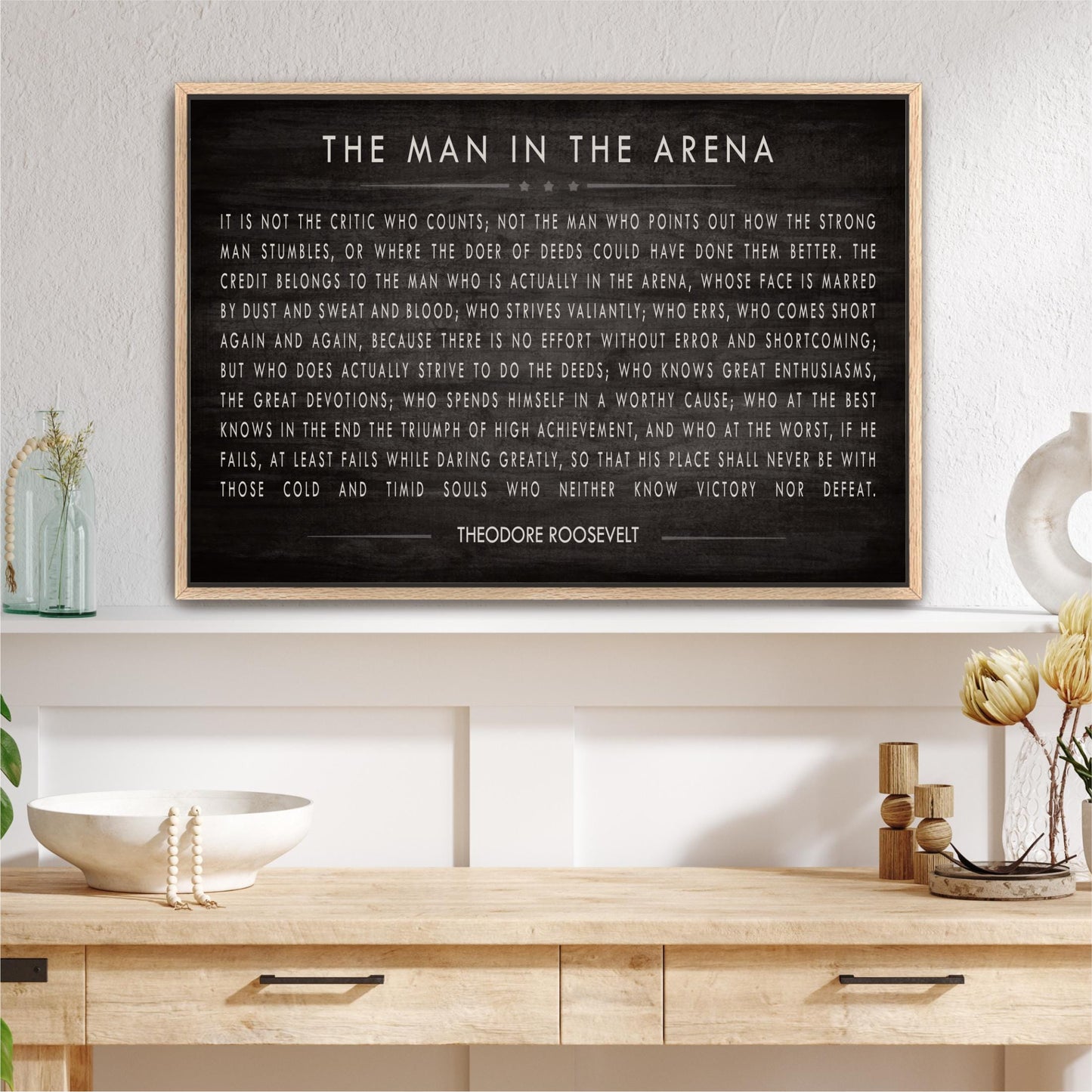 a framed quote by Theodore Roosevelt, "The Man in the Arena", displayed on a wall above a wooden shelf with various decorative items such as a vase, a bowl, and a potted plant.