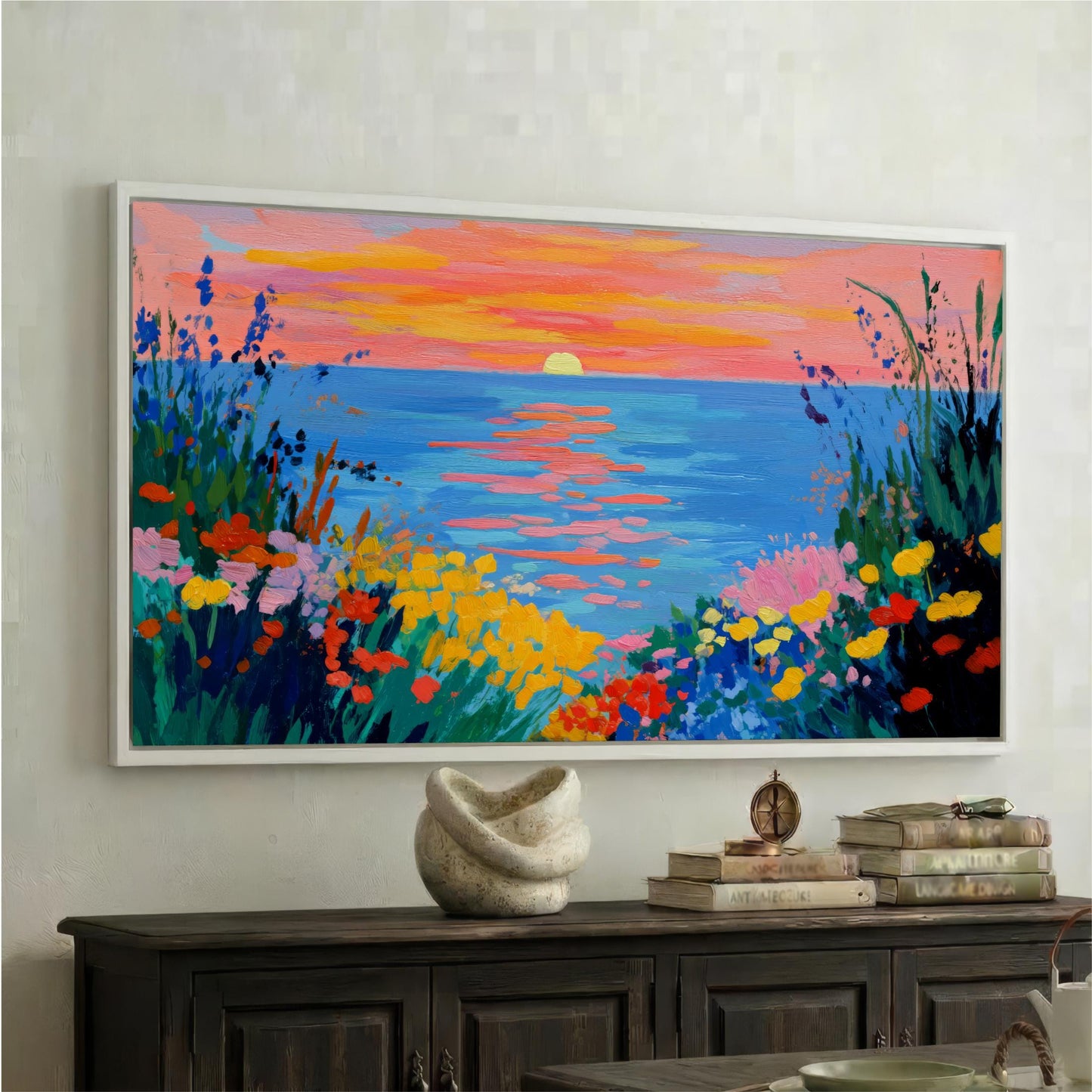 a large, colorful painting of a sunset over a body of water, with a vibrant display of orange, pink, and blue hues. The painting is framed and hung on a wall above a wooden cabinet, which has a white vase and a stack of books on top.