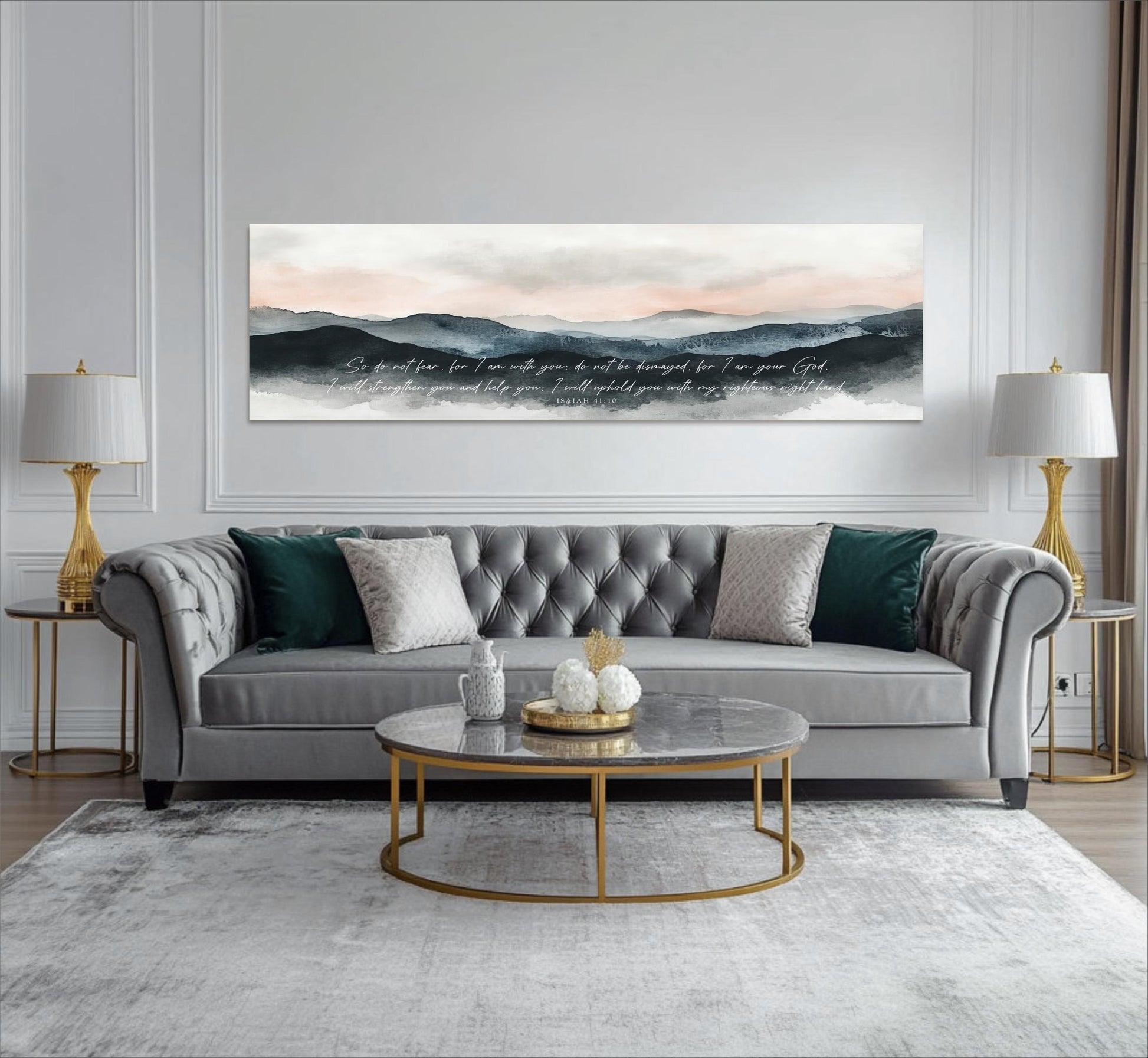 a modern living room with a gray tufted sofa, a round marble coffee table, and two gold-framed lamps on either side of the sofa. Above the sofa, there is a large canvas print depicting a mountain landscape with a quote written in cursive font.