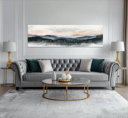 a modern living room with a gray tufted sofa, a round marble coffee table, and two gold-framed lamps on either side of the sofa. Above the sofa, there is a large canvas print depicting a mountain landscape with a quote written in cursive font.