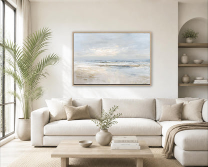 a serene living room with a large painting of a beach scene hanging on the wall above a white sofa. The room is decorated with a potted plant, a vase with greenery, and a stack of books on a coffee table.