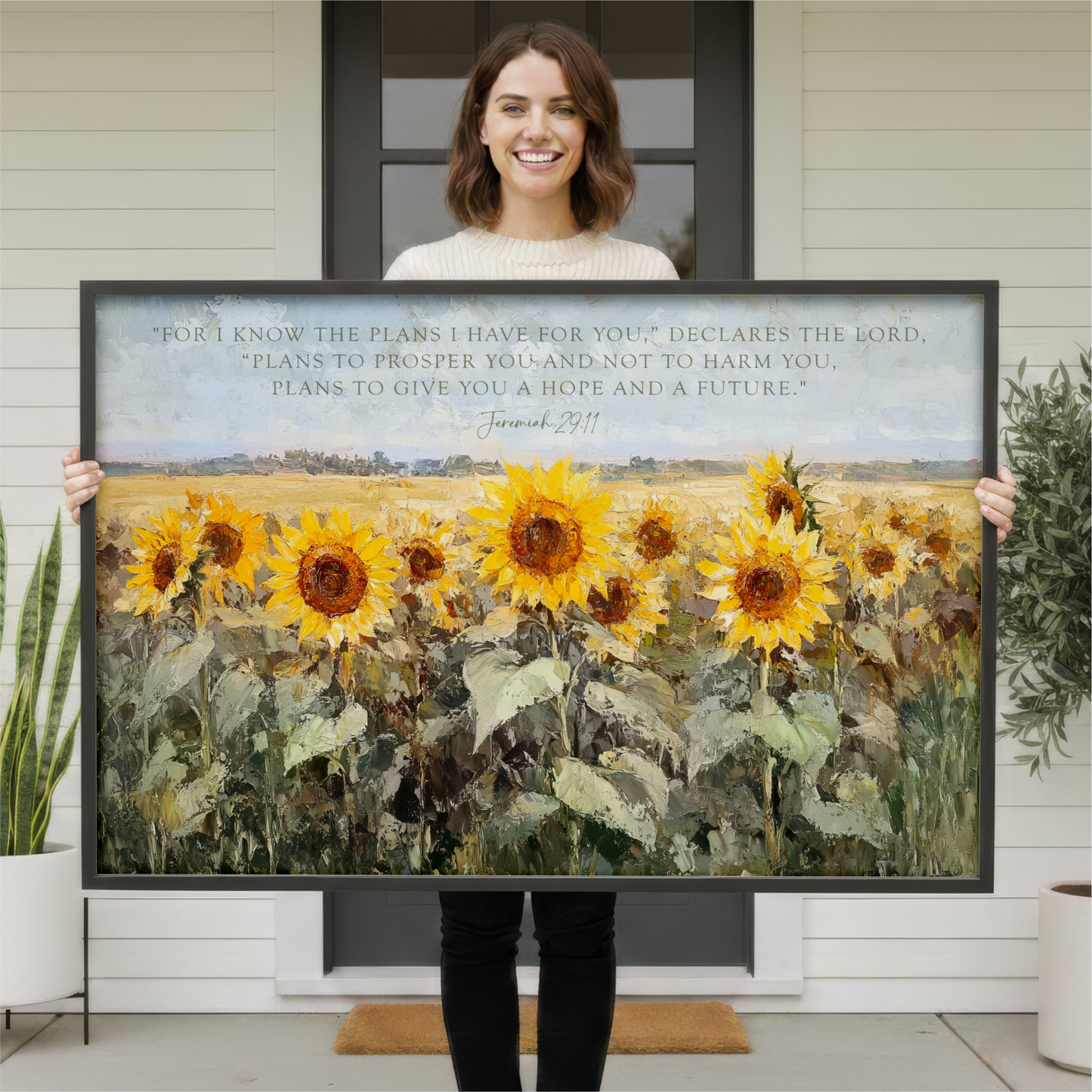 A woman is holding up a large painting of a field of sunflowers in front of a house.