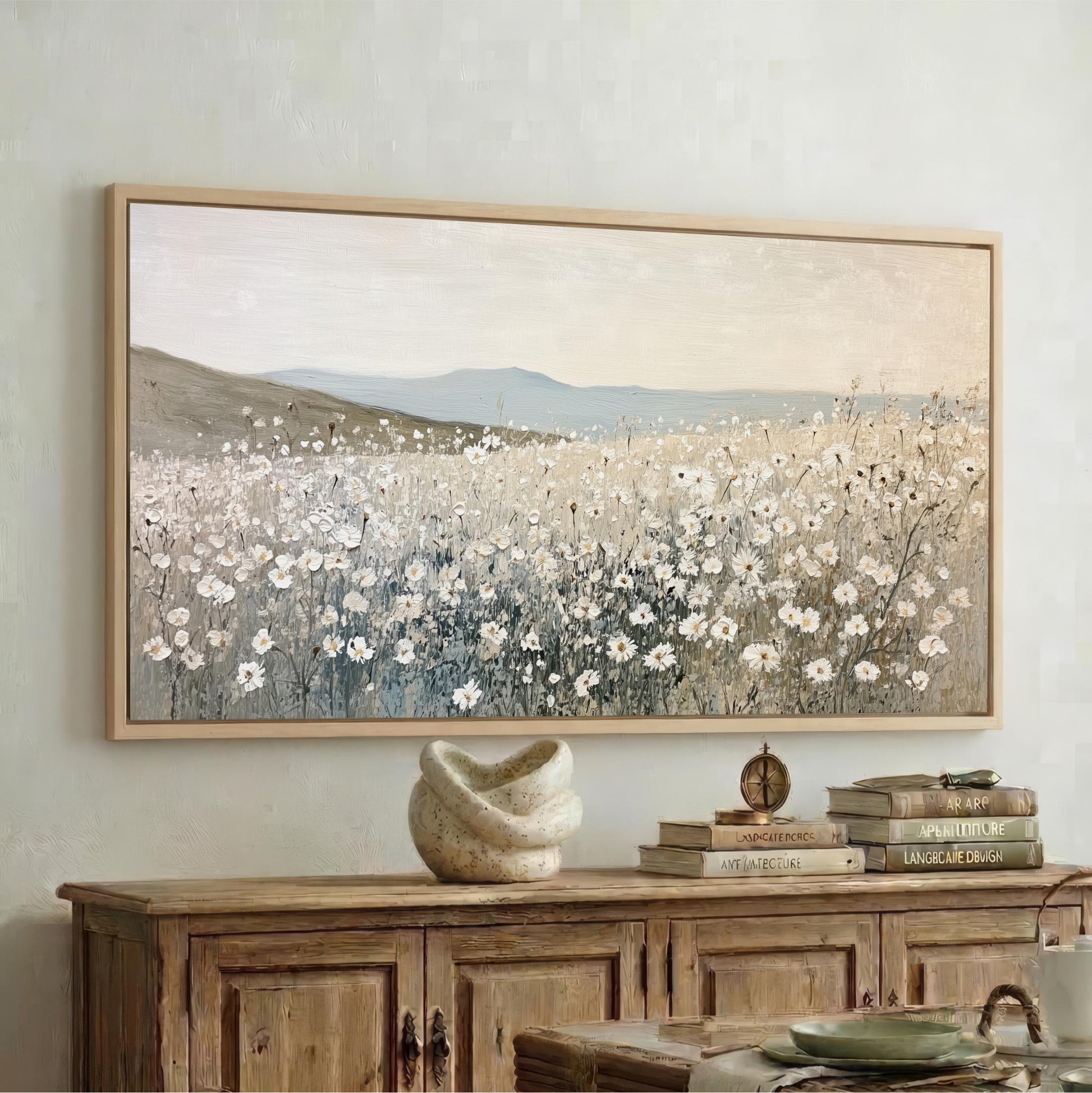 a large painting of a field of flowers hanging on a wall above a wooden cabinet. The cabinet has a stack of books and a decorative vase on top of it.
