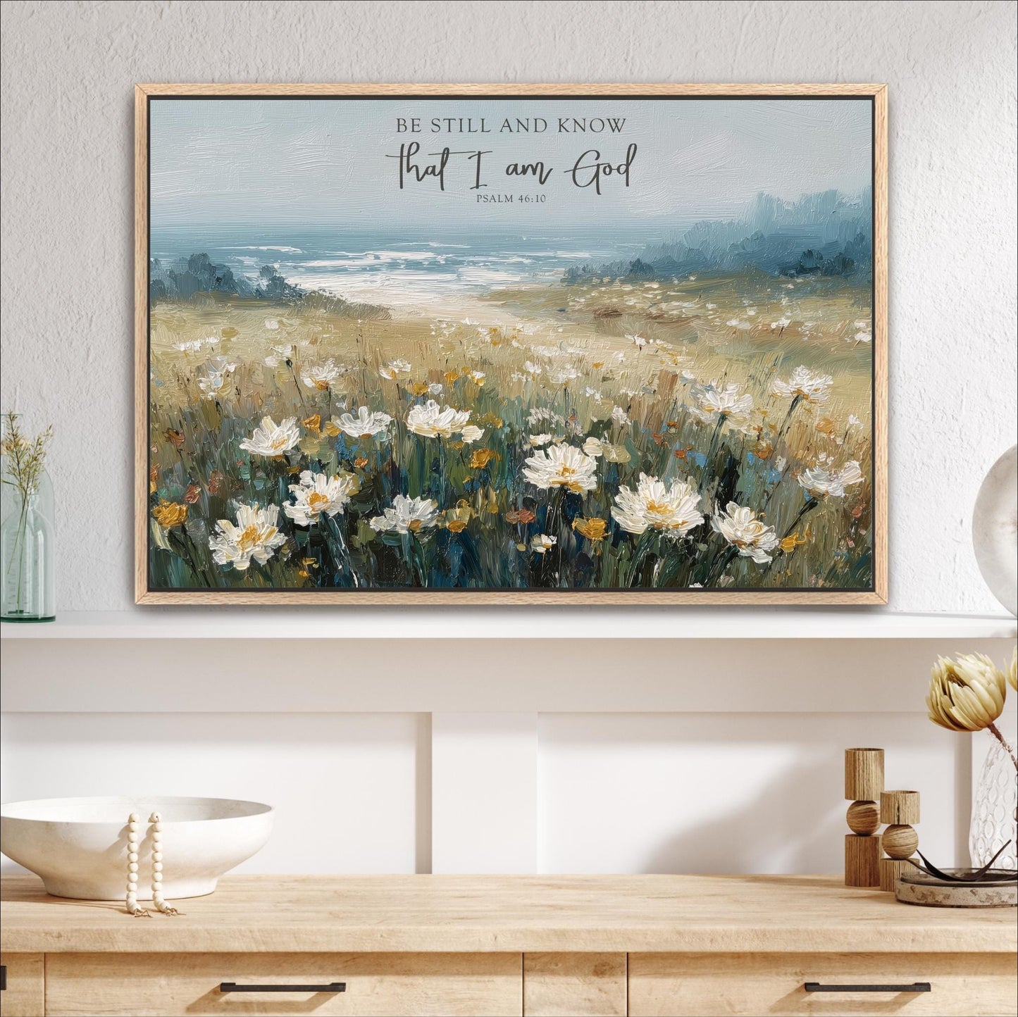a framed painting of a field of white flowers, with a blue sky and water in the background. The painting is hung on a wall above a wooden shelf.