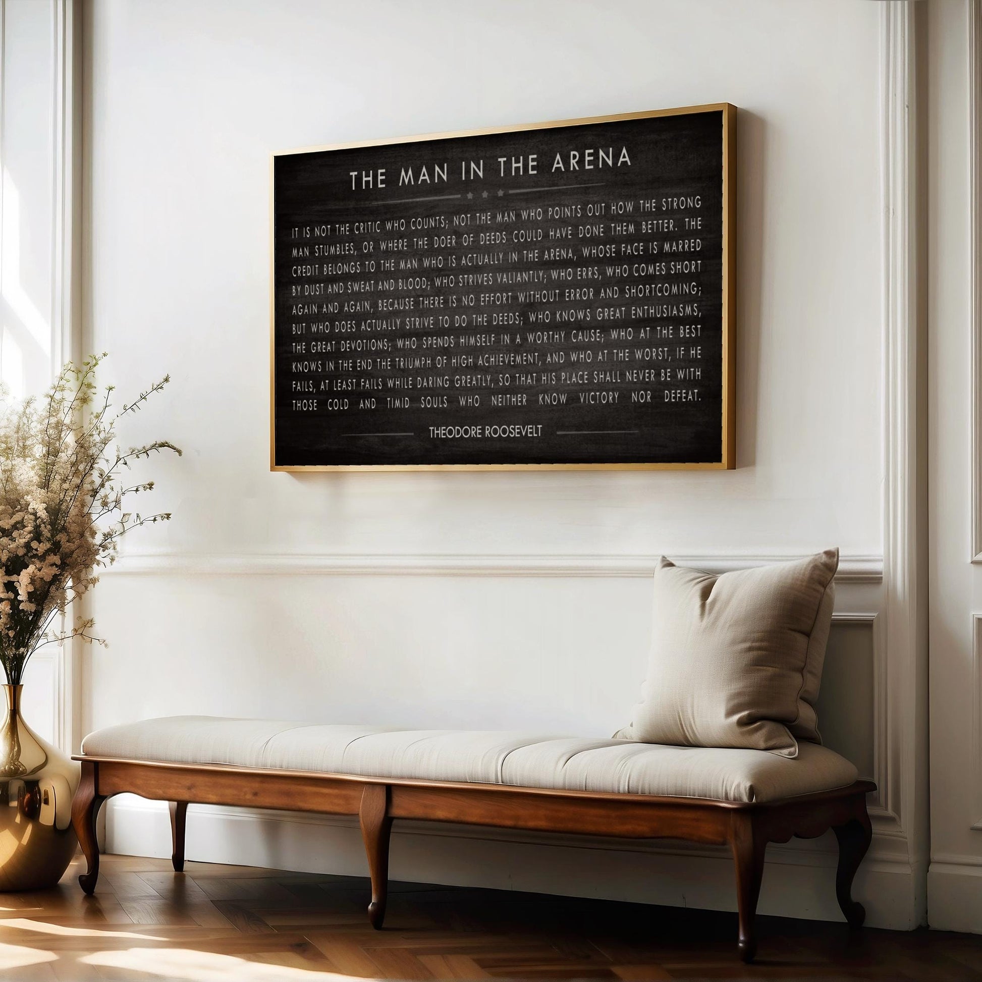 a framed quote by President Franklin D. Roosevelt, which reads "The man in the arena" and is displayed on a white wall above a wooden bench.