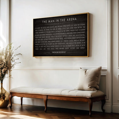 a framed quote by President Franklin D. Roosevelt, which reads "The man in the arena" and is displayed on a white wall above a wooden bench.