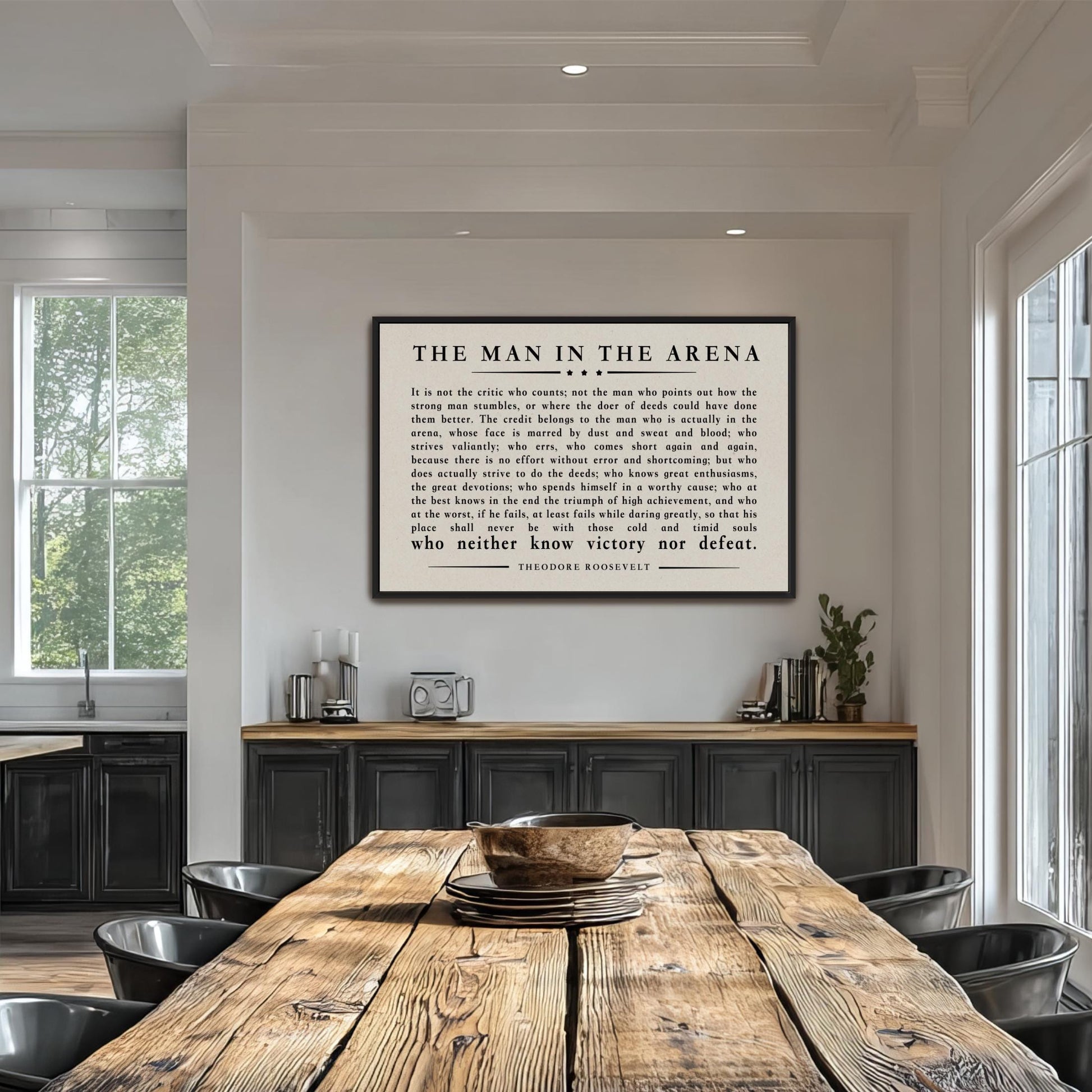 a wooden dining table with a bowl on it, surrounded by chairs. Above the table, there is a framed quote in black text that reads "The man in the arena".