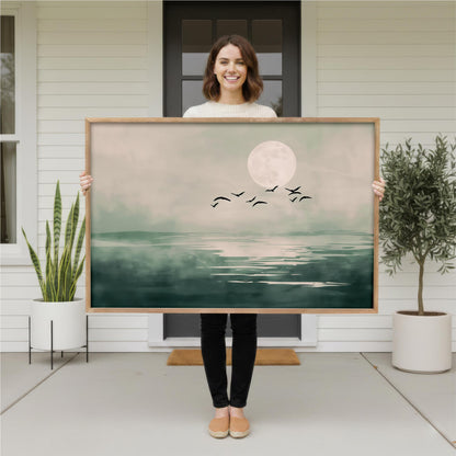 A woman is holding a large framed painting of a serene landscape with a full moon, birds, and a body of water.