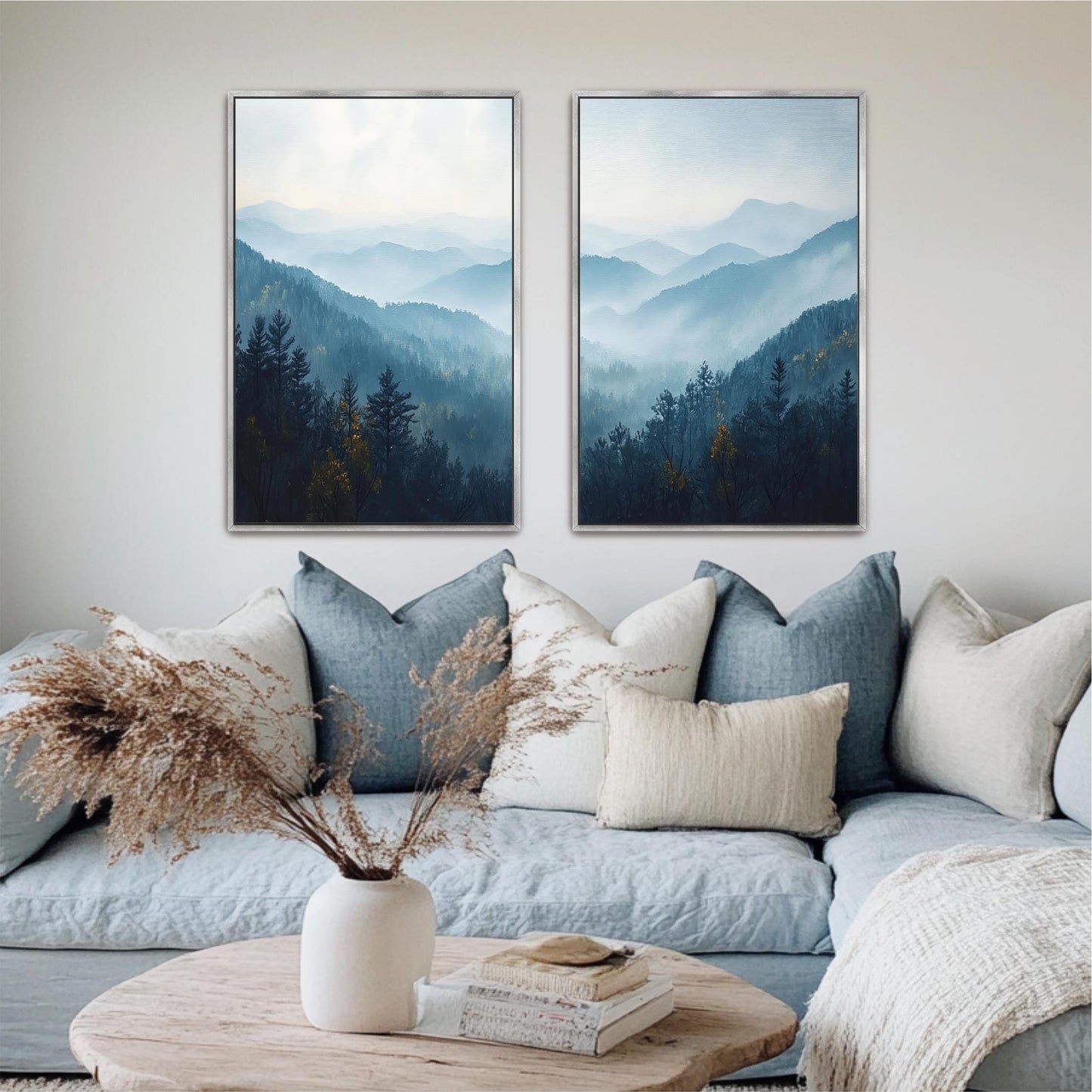 a cozy living room with a blue couch adorned with white and beige pillows, a white vase with dried flowers, and two framed landscape paintings hanging on the wall above the couch.