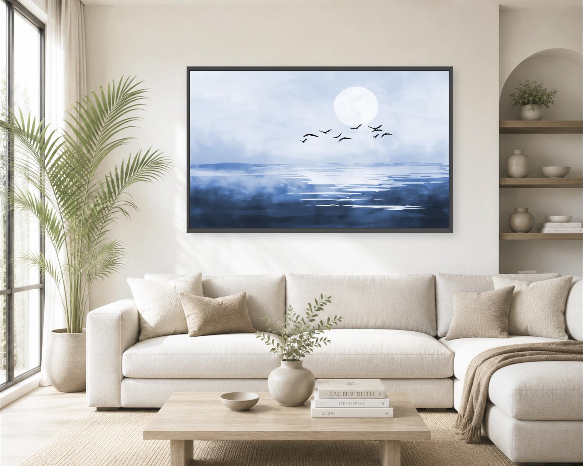 a serene living room with a large painting of a blue sky and white moon hanging on the wall above a white sofa. The room is decorated with a potted plant, a vase with greenery, and a stack of books on a coffee table.