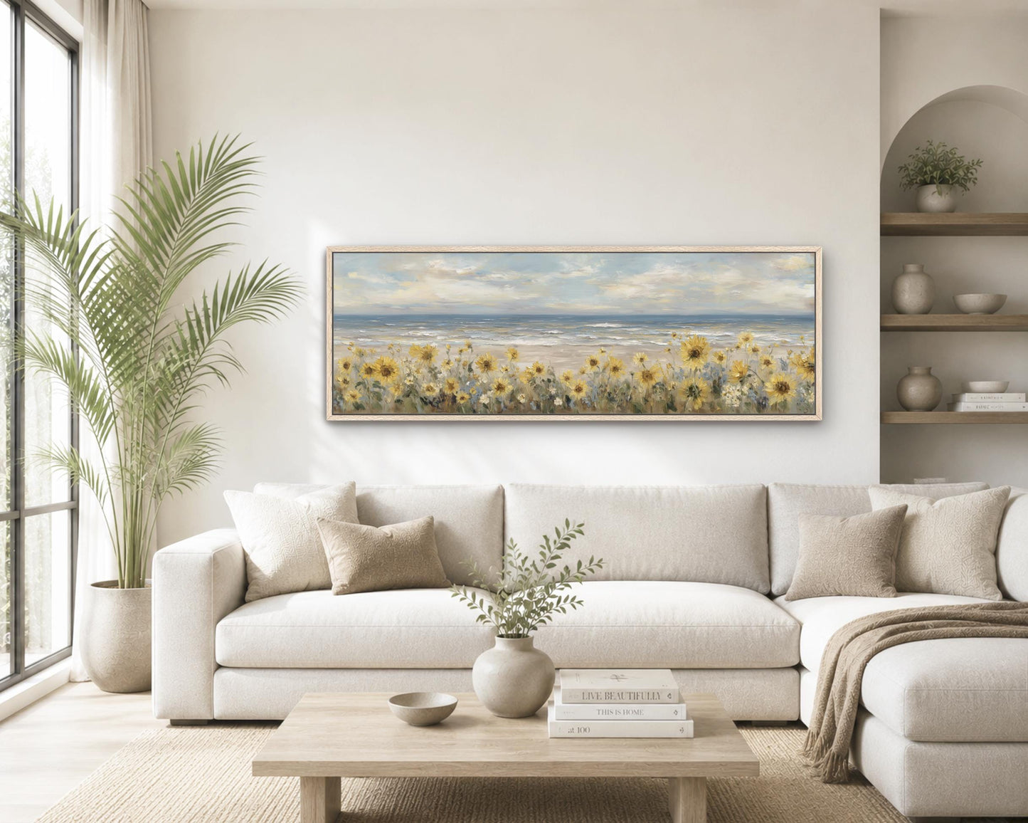 a serene living room with a large painting of a field of sunflowers hanging on the wall above a white sofa. The sofa is positioned in front of a wooden coffee table, which has a vase of greenery and a stack of books on it. The room also features a large window, a potted plant, and a bookshelf with various decorative items.