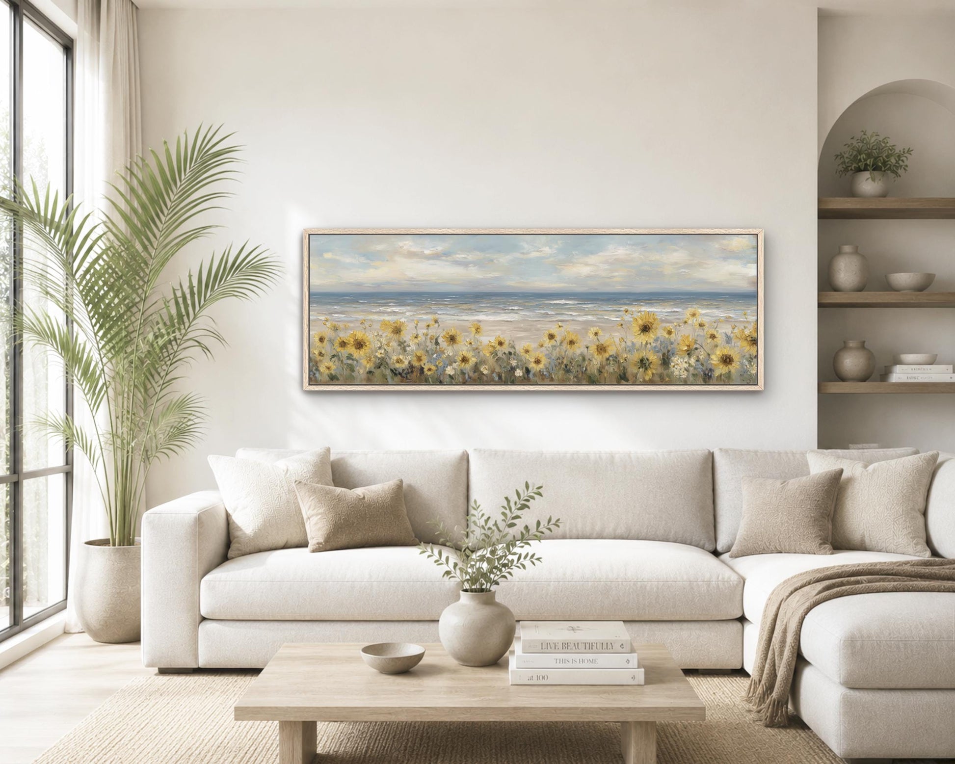 a serene living room with a large painting of a field of sunflowers hanging on the wall above a white sofa. The sofa is positioned in front of a wooden coffee table, which has a vase of greenery and a stack of books on it. The room also features a large window, a potted plant, and a bookshelf with various decorative items.