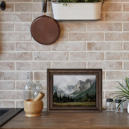 a framed landscape painting hanging on a brick wall above a wooden table with various kitchen items, including a wooden bowl, a mortar and pestle, and a plant.