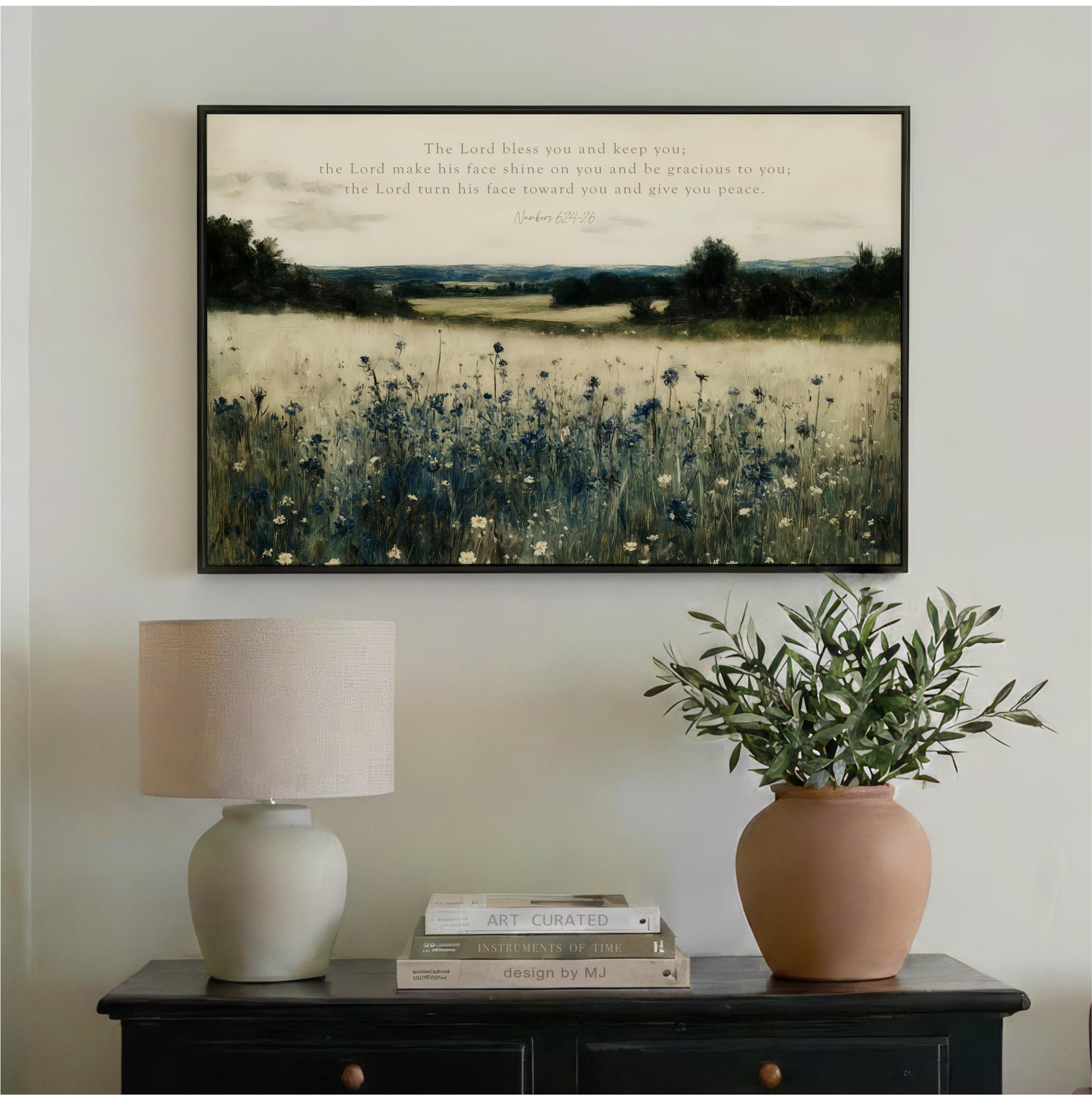 a framed painting of a field with wildflowers, a lamp, and a potted plant on a wooden table.