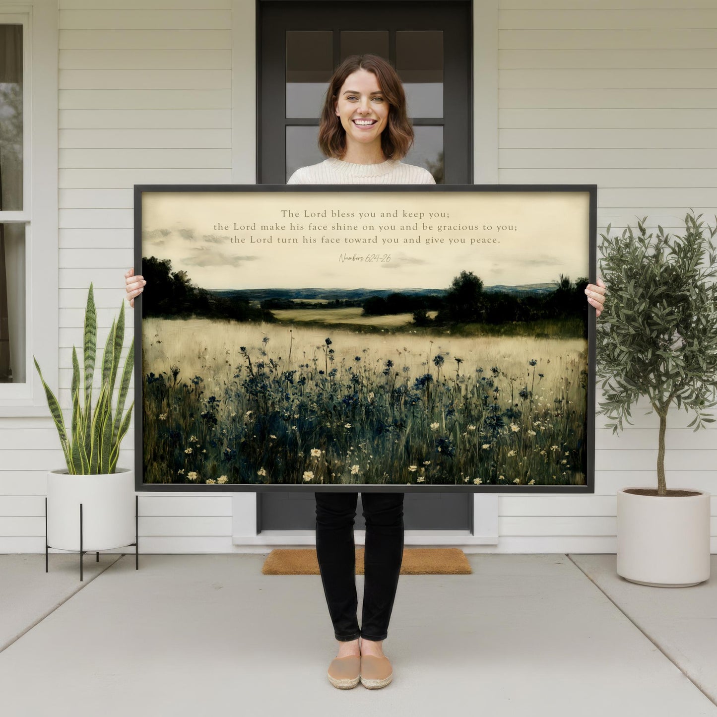 A woman stands in front of a white house, holding a large framed print of a serene landscape with a field of flowers and a house in the background.