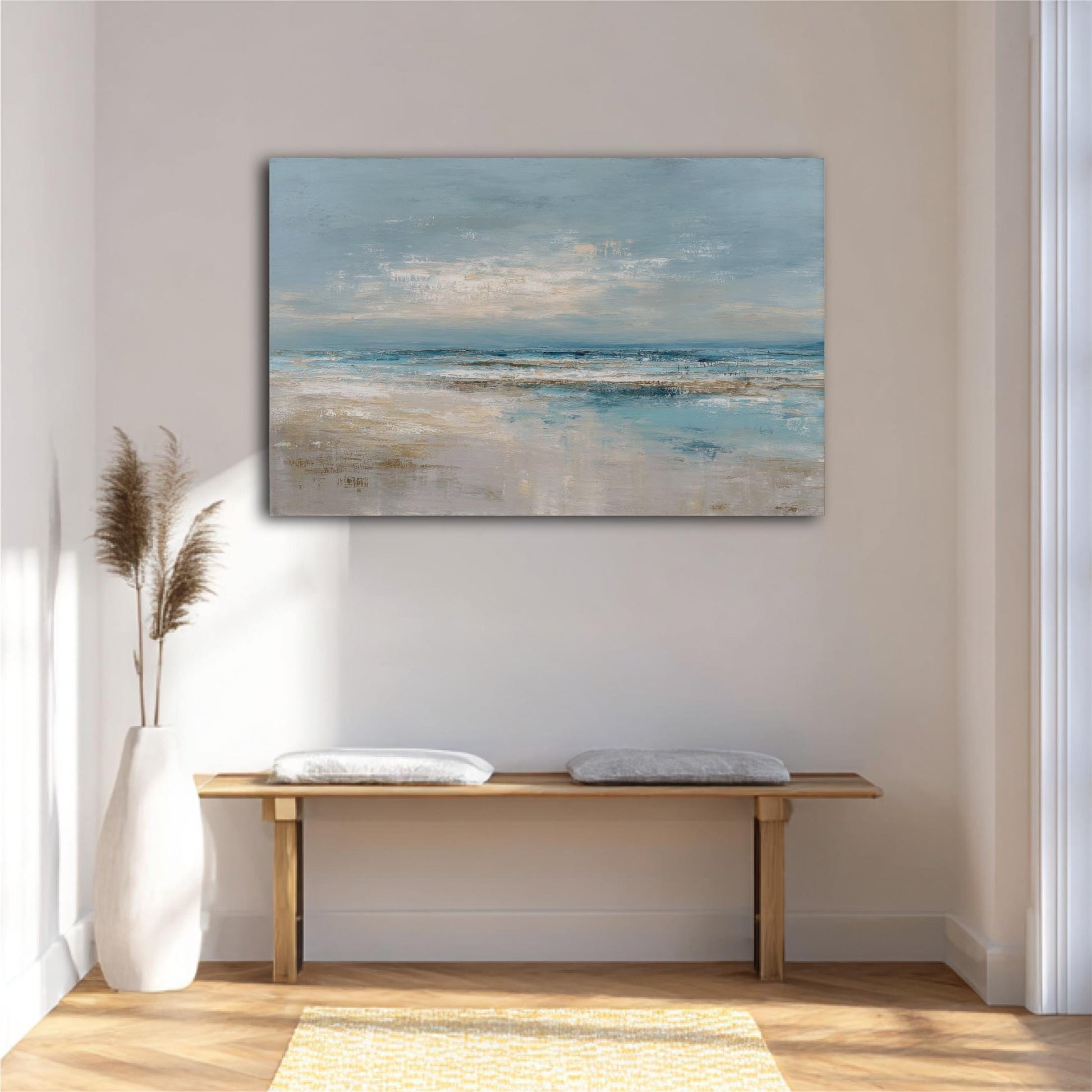 a serene indoor setting with a large painting of a beach scene hanging on the wall above a wooden bench with two white cushions. The painting depicts a tranquil beach landscape with a blue sky, white clouds, and a sandy shoreline. The room has a minimalist and modern design, with a white vase and a yellow rug adding to the overall aesthetic.