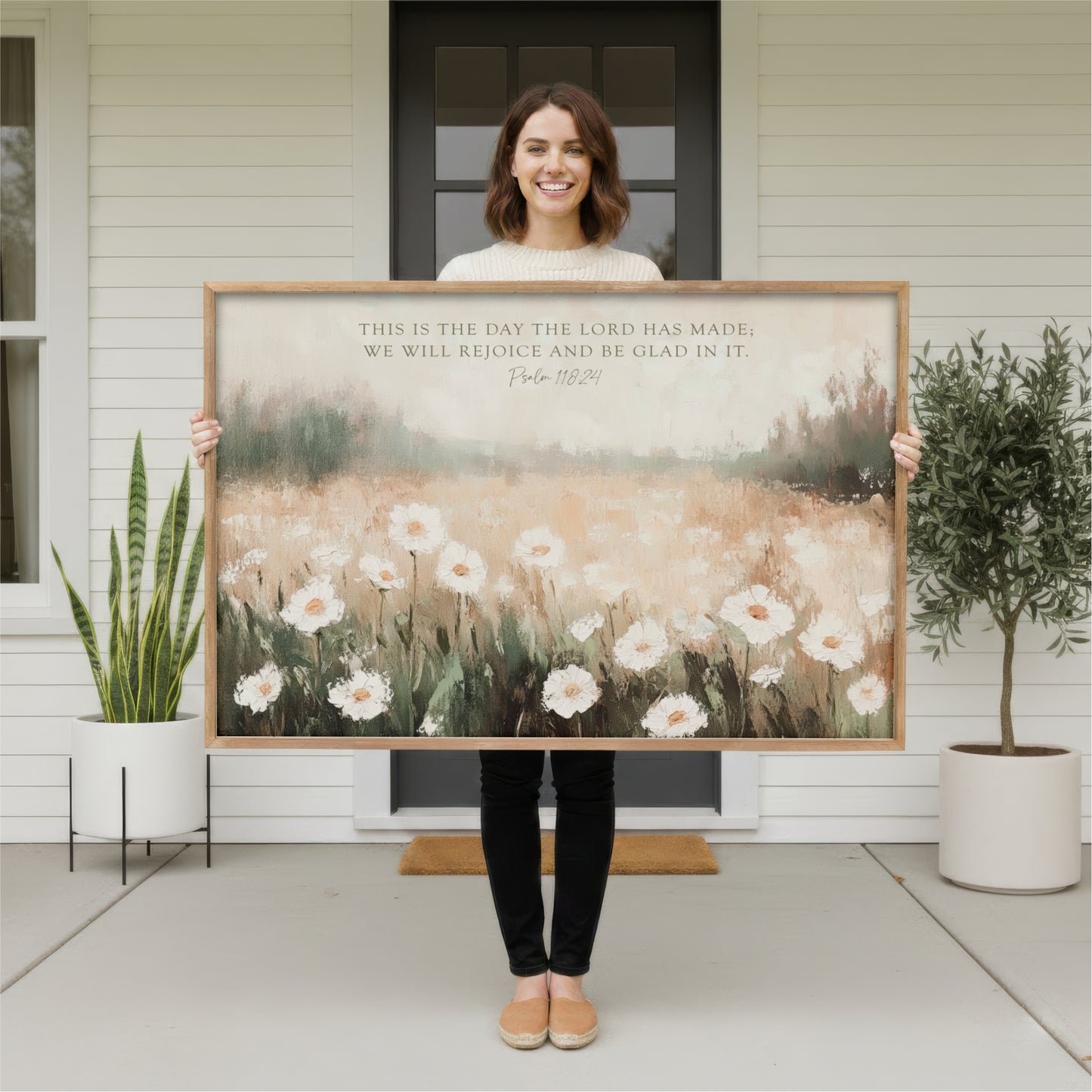 A woman stands in front of a white house, holding a large painting of a field of white flowers.