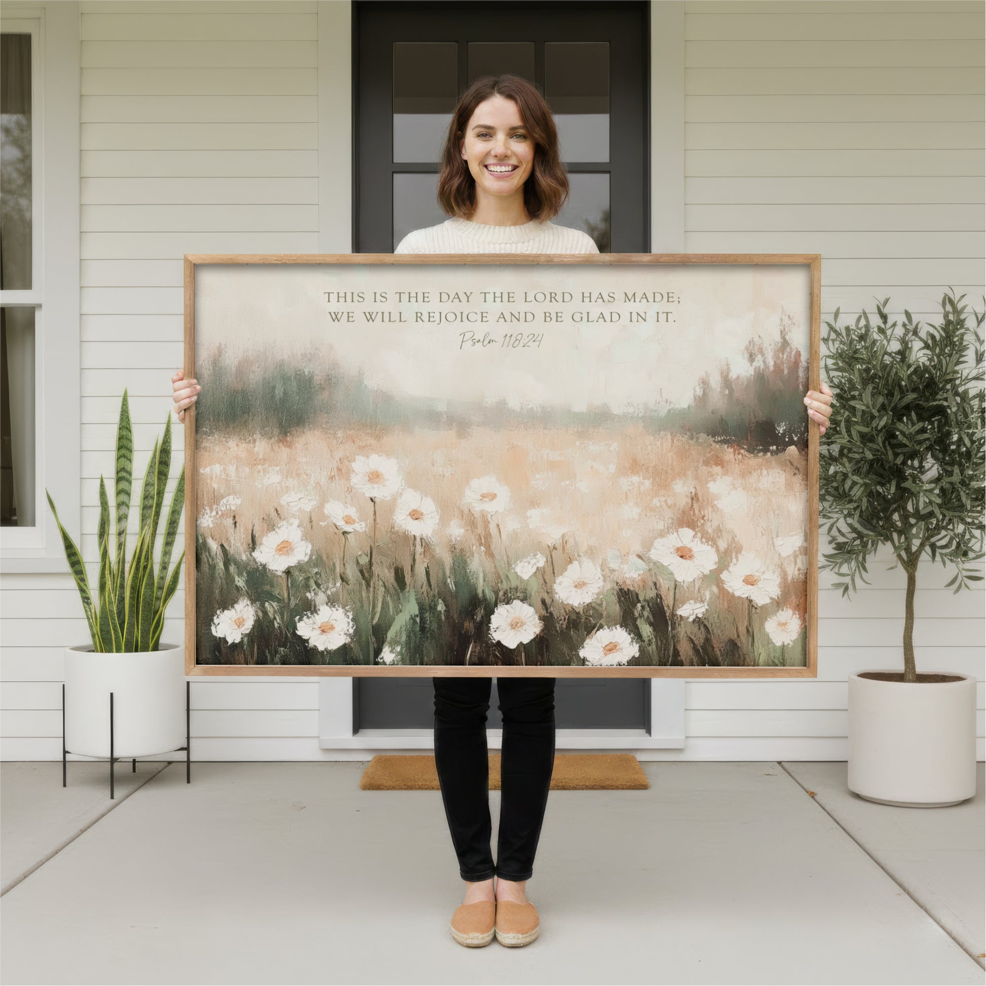 A woman stands in front of a white house, holding a large painting of a field of white flowers.