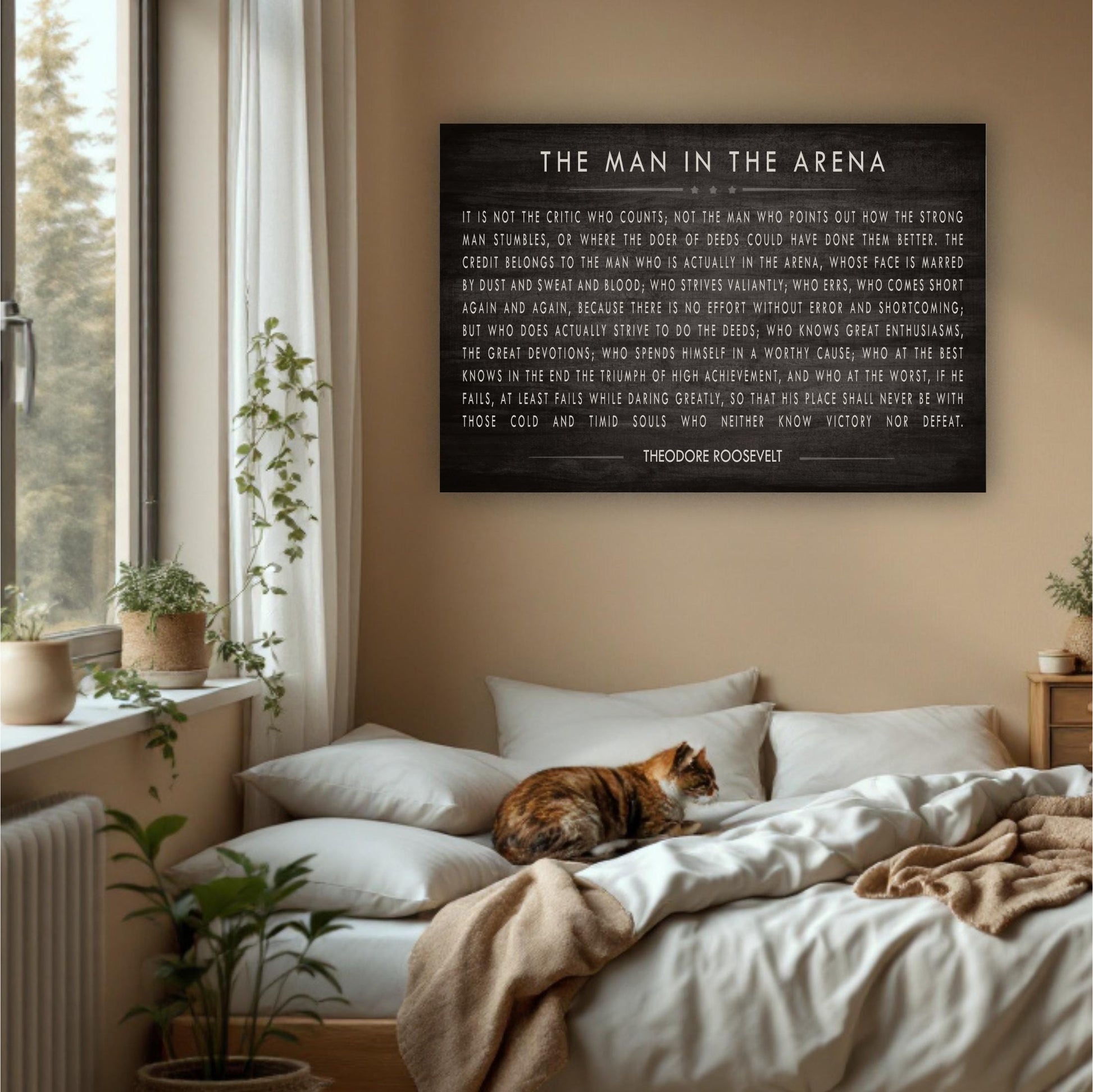 a bedroom with a large black sign hanging on the wall that reads "The Man in the Arena" by Theodore Roosevelt. The sign is surrounded by various objects, including a cat sleeping on the bed, potted plants, and a window.