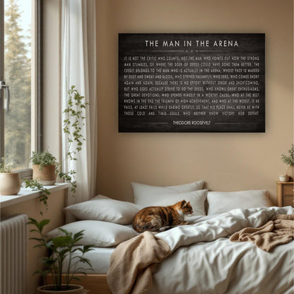 a bedroom with a large black sign hanging on the wall that reads "The Man in the Arena" by Theodore Roosevelt. The sign is surrounded by various objects, including a cat sleeping on the bed, potted plants, and a window.