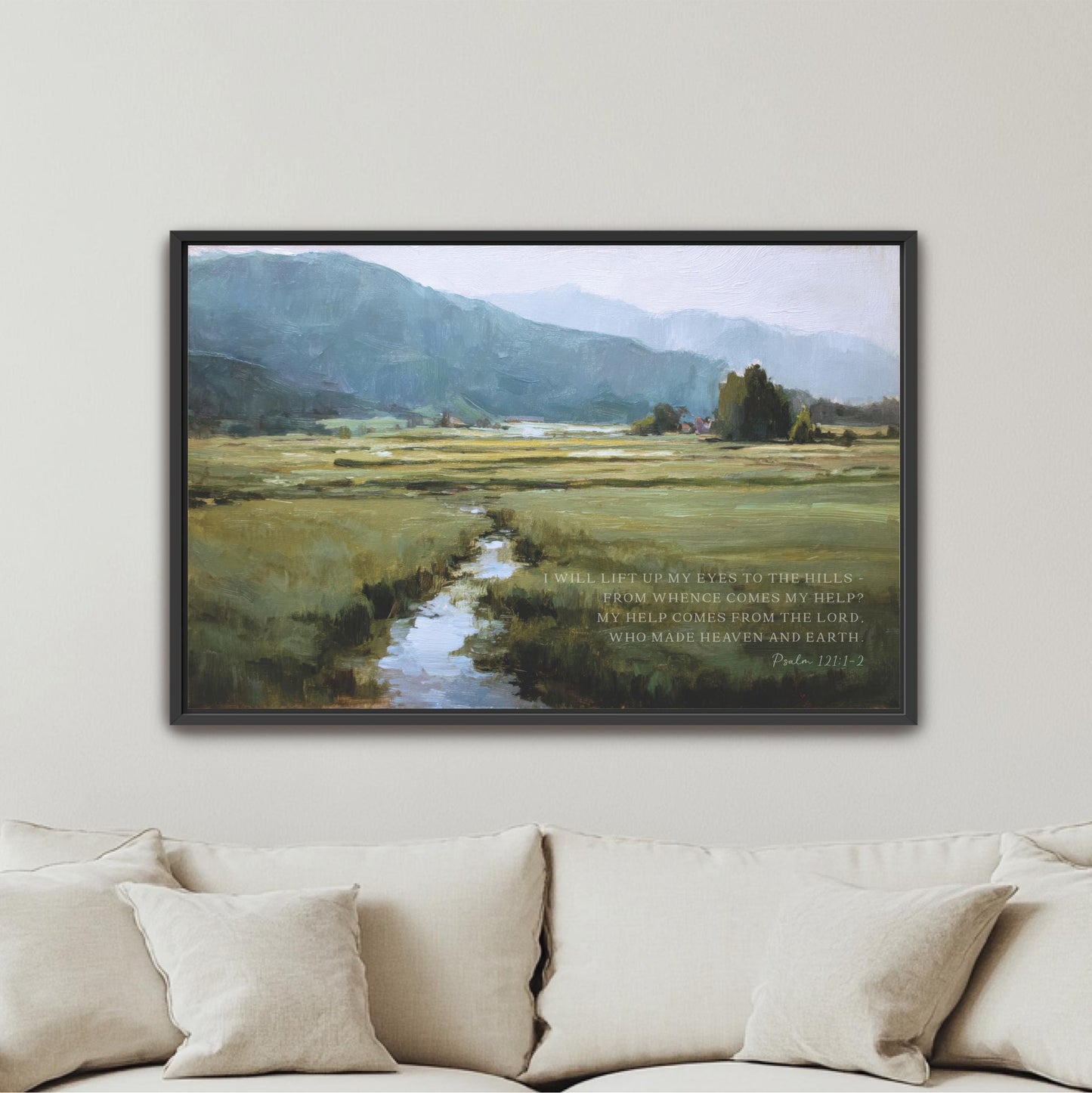 The image is a framed painting of a serene landscape with a winding stream, surrounded by mountains and lush greenery. The painting is hung on a wall above a beige couch, creating a peaceful and inviting atmosphere in the room.