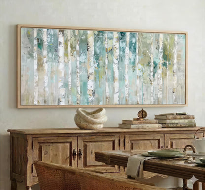 a large, abstract painting of trees with a mix of green, blue, and white colors, hanging on a wall above a wooden cabinet. The cabinet has a vase and some books on top of it. In the foreground, there is a wooden table with a bowl and a plate on it, and a woven chair is visible.