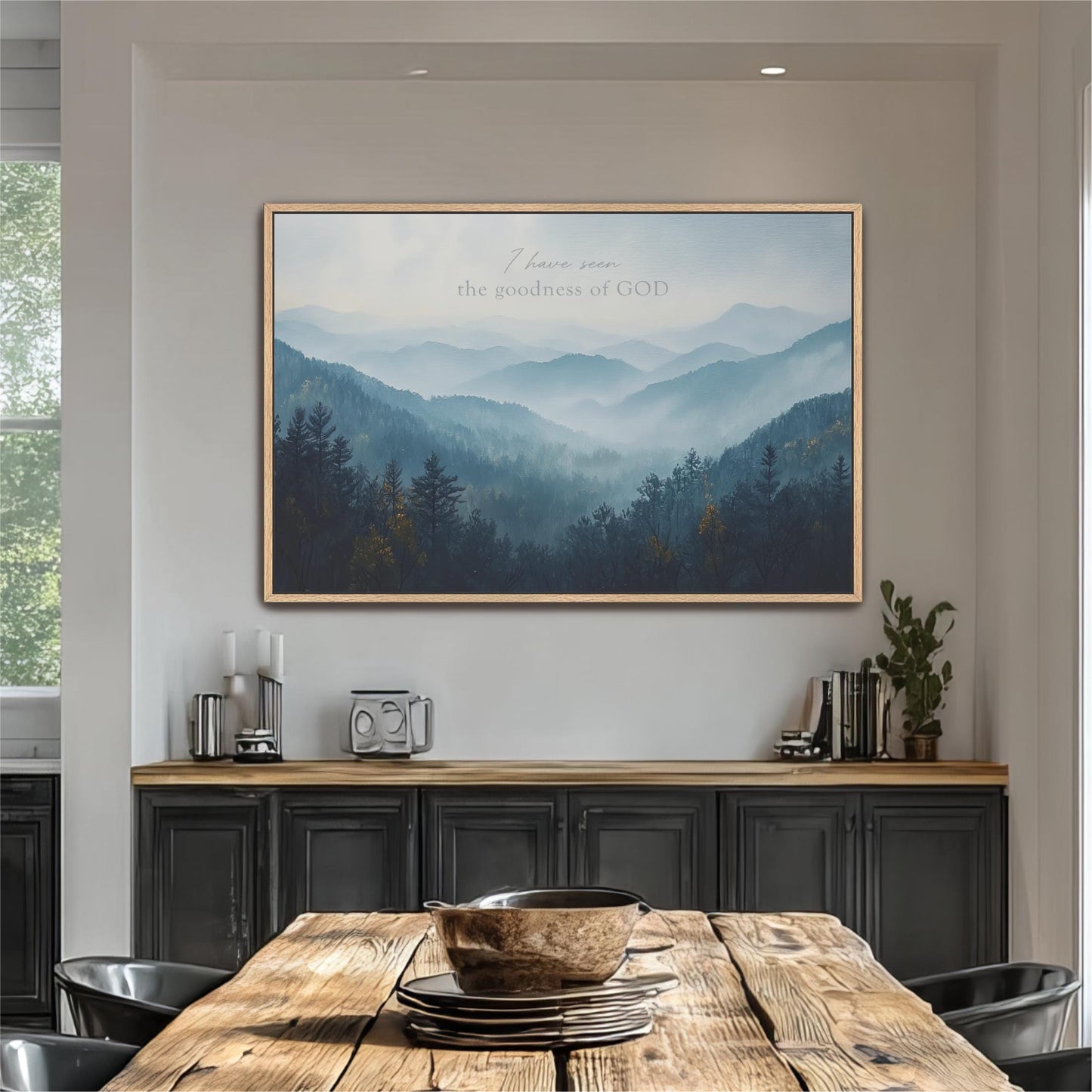 a large framed picture of a mountain landscape hanging on a wall above a wooden dining table with a bowl on it.
