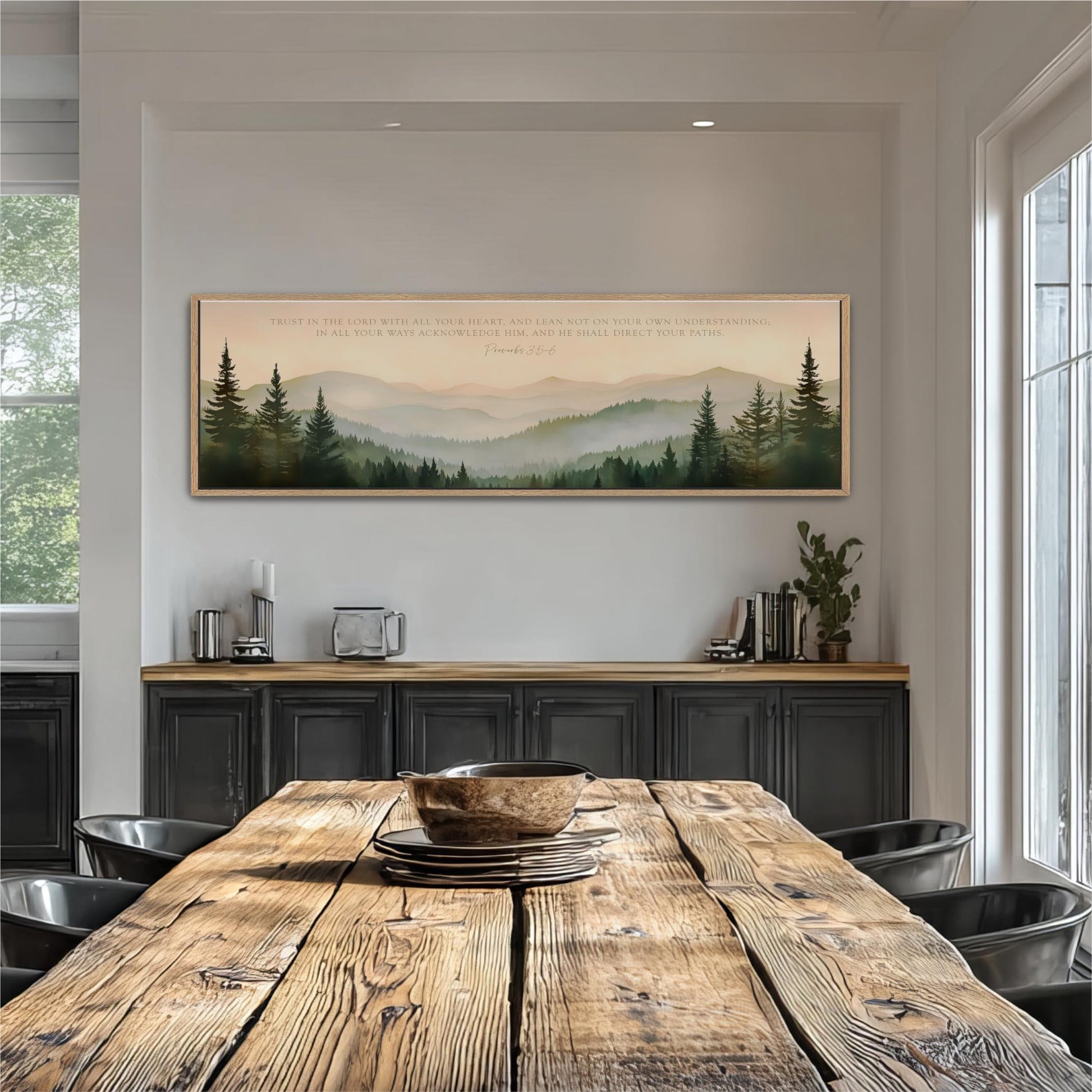 a rustic wooden dining table with a bowl on top, surrounded by black chairs. Above the table, there is a large framed picture depicting a mountain landscape with pine trees. The wall behind the table has a shelf with various items, including a potted plant and a vase.