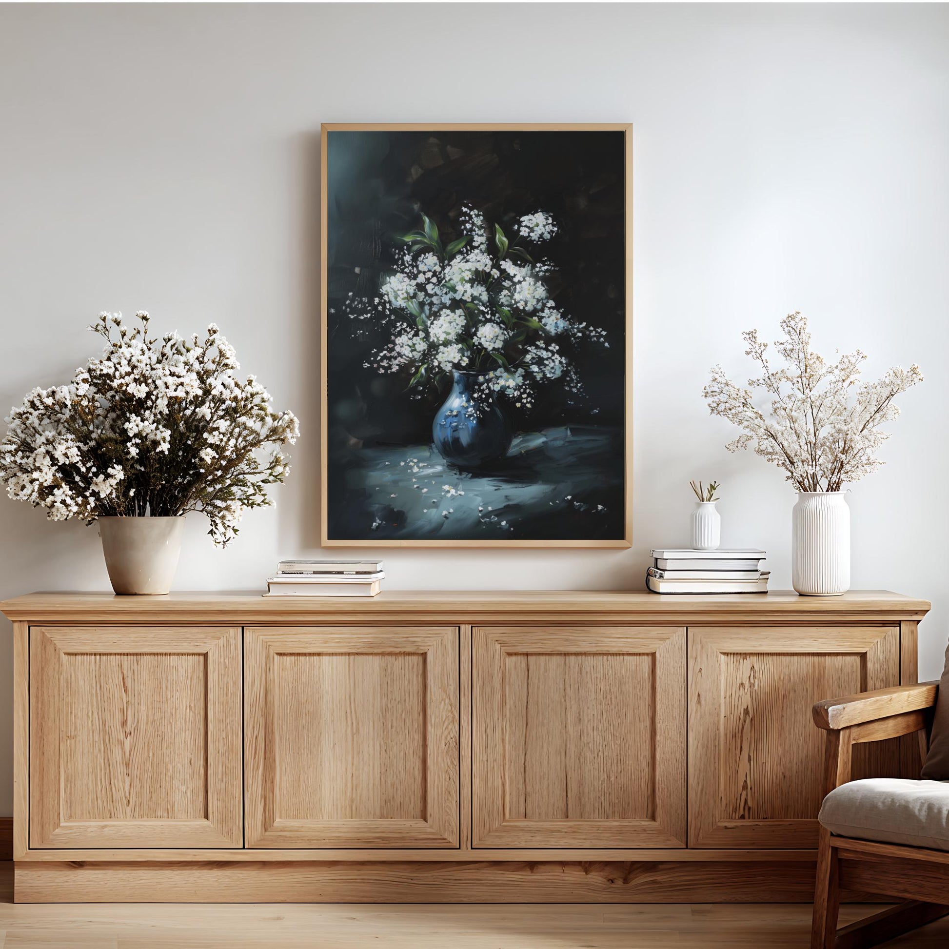 a wooden cabinet with a framed painting of a vase filled with white flowers hanging above it. The cabinet has three doors and is situated in a room with a white wall and a wooden floor. There are also two vases with white flowers placed on the cabinet, and a stack of books is visible on the right side of the cabinet.