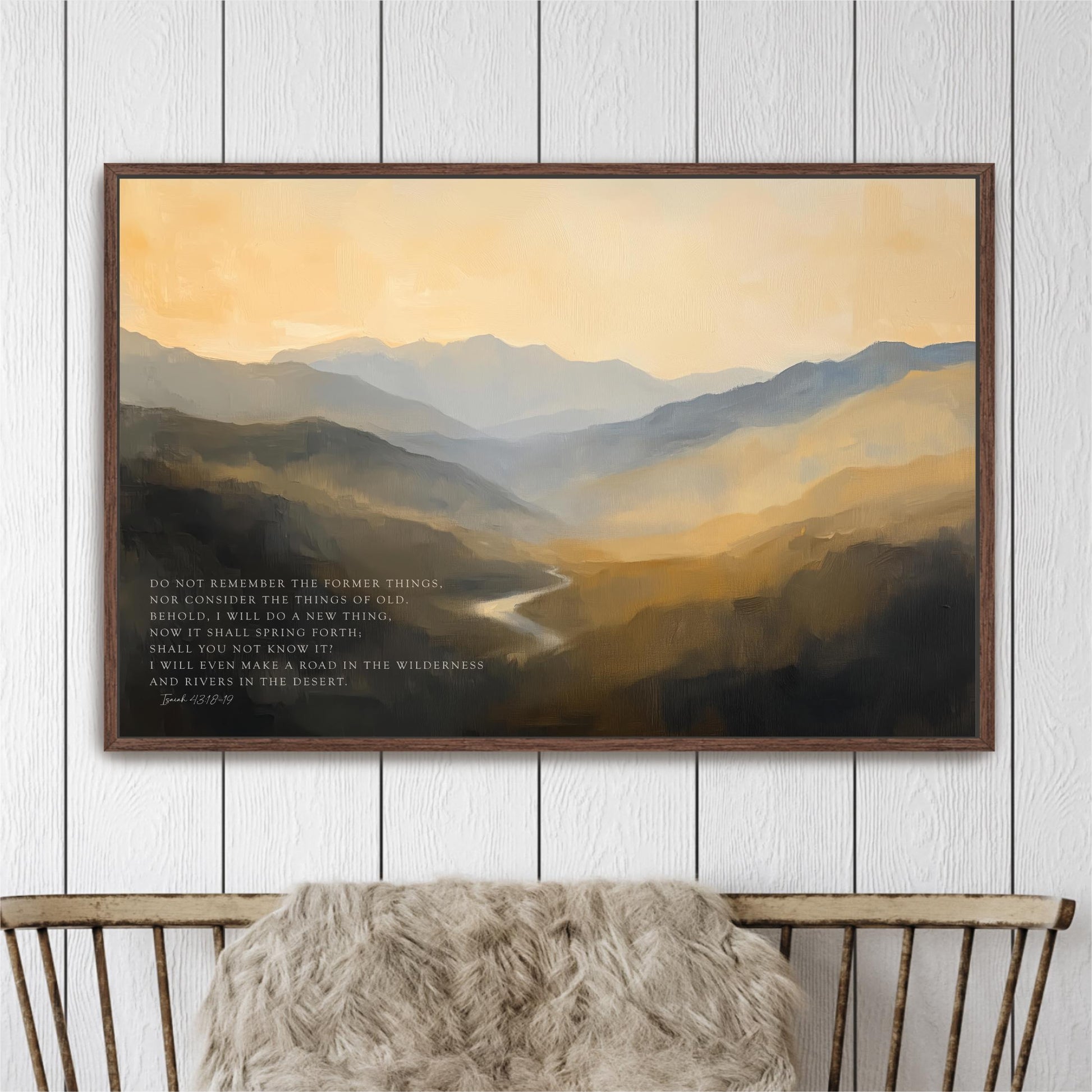 a framed painting of a mountainous landscape with a river winding through it, hanging on a white wall above a wooden bench.