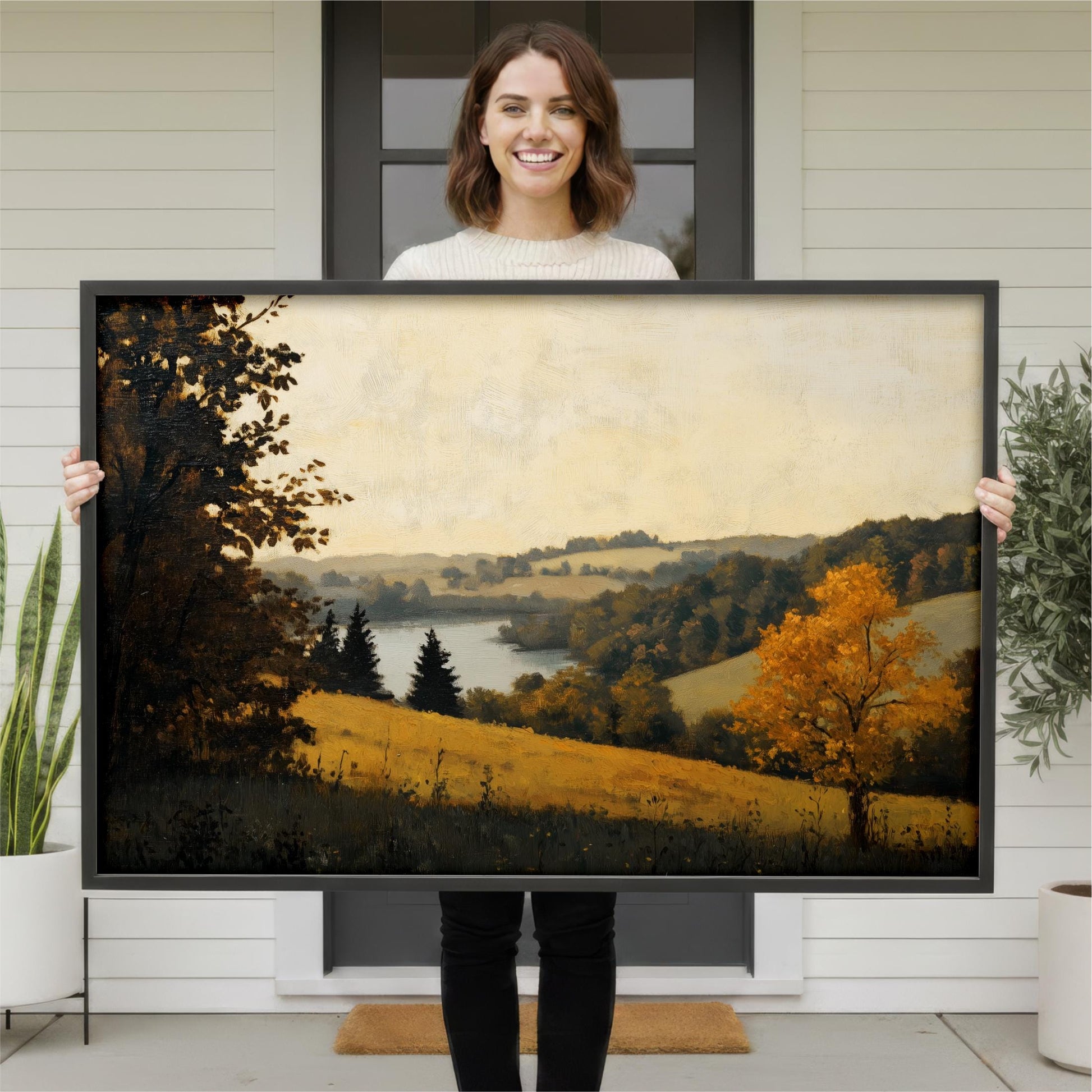 A woman is holding a large framed painting of a countryside landscape, which features a field, trees, and a body of water.