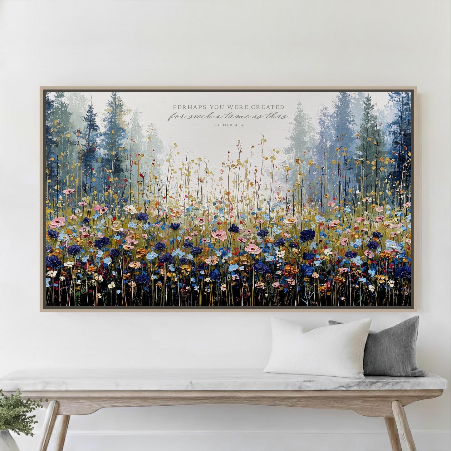 The image is a large framed painting depicting a vibrant field of flowers, with a forest in the background. The painting is hung on a wall above a wooden bench, which has two pillows on it.