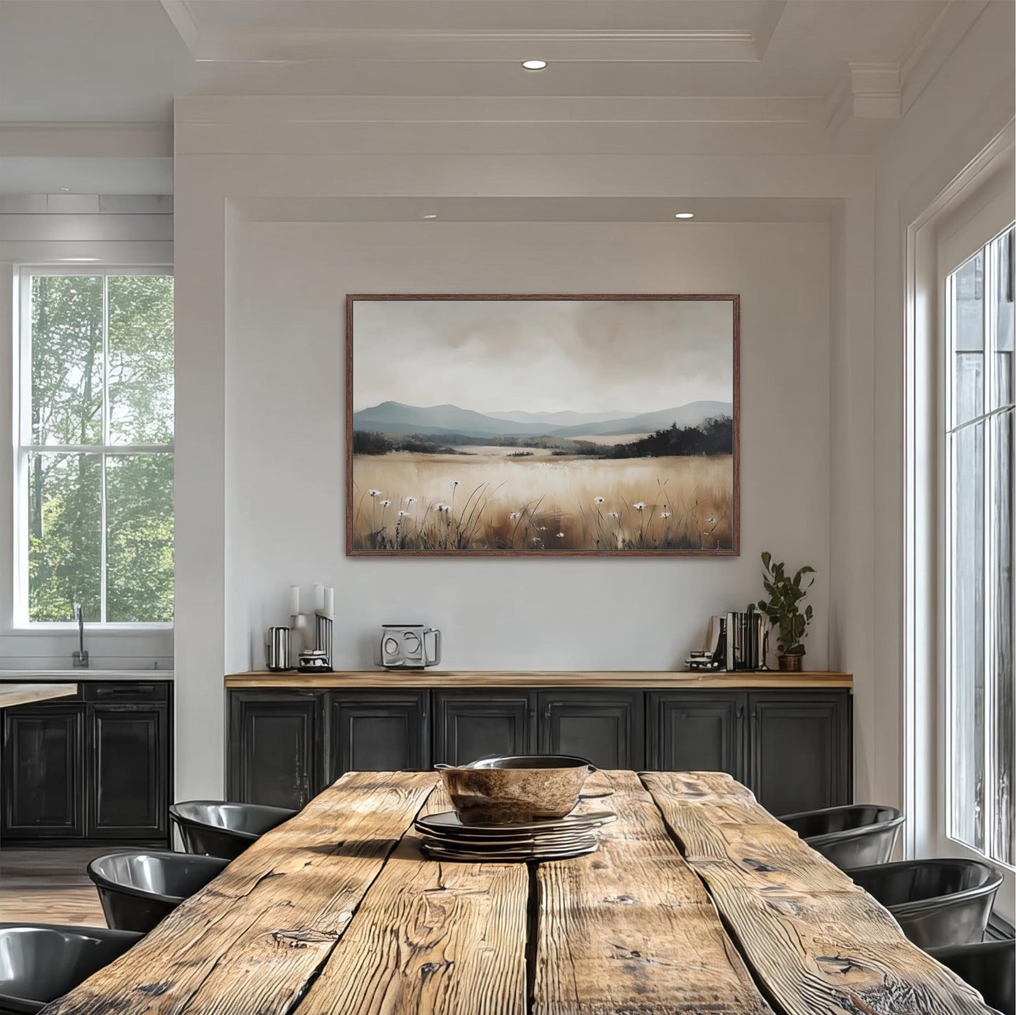 a dining room with a large wooden table surrounded by black chairs. Above the table, there is a framed painting depicting a serene landscape with a field and mountains in the background. The room has white walls and a window that allows natural light to enter the space.