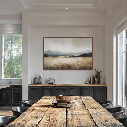 a dining room with a large wooden table surrounded by black chairs. Above the table, there is a framed painting depicting a serene landscape with a field and mountains in the background. The room has white walls and a window that allows natural light to enter the space.