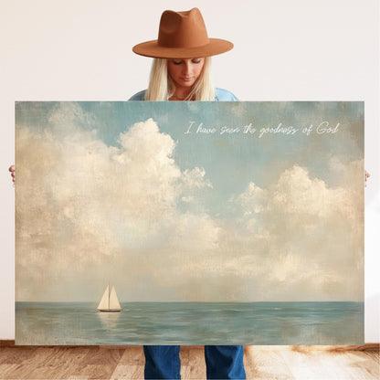 A woman in a brown hat holds a large canvas painting depicting a sailboat on the ocean, with the text "I have seen the goodness of God" written above the painting.