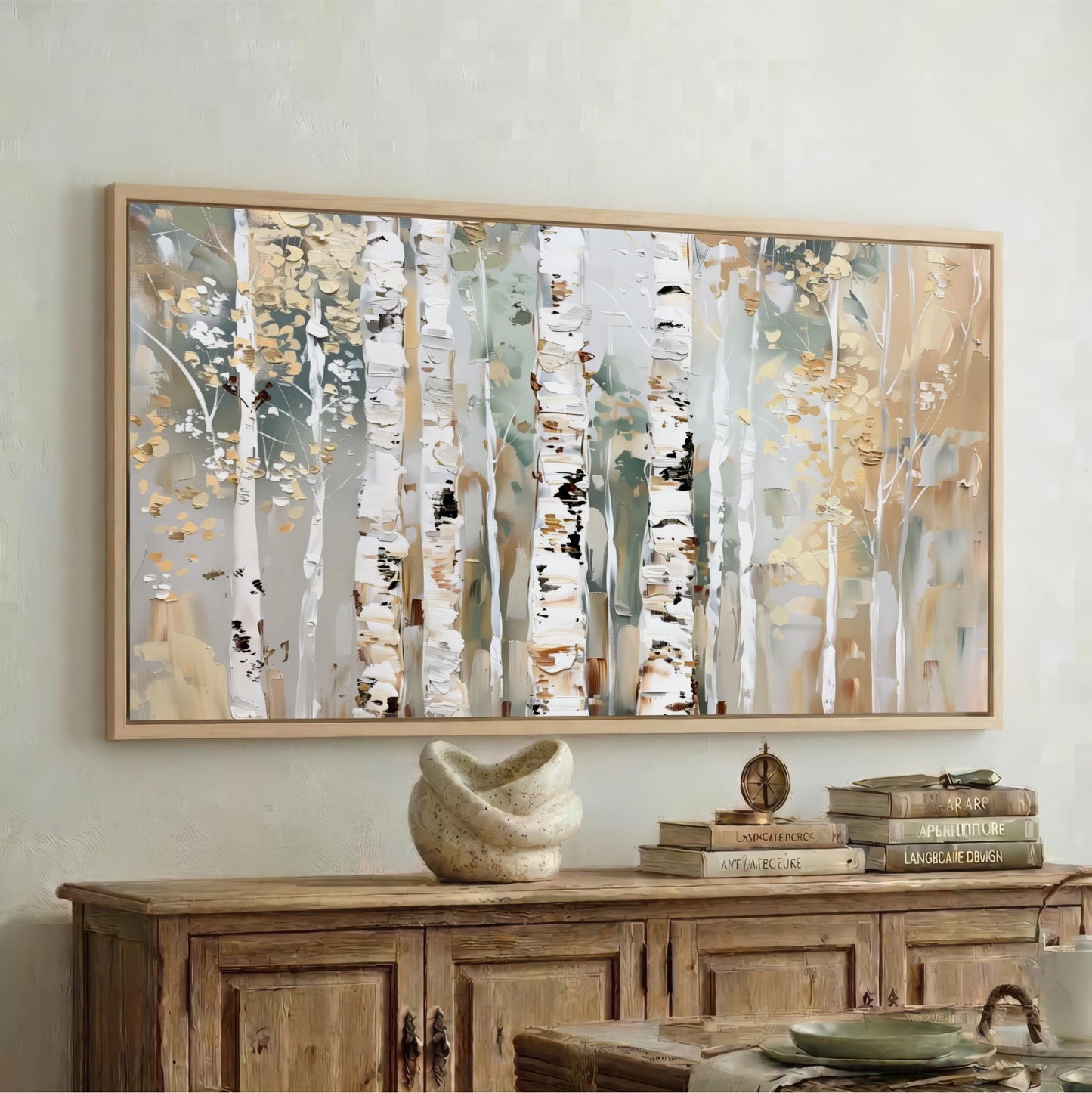 a large framed painting of birch trees with a white and gold color scheme, hanging on a wall above a wooden cabinet. The cabinet has a stack of books and a small white sculpture on top of it.
