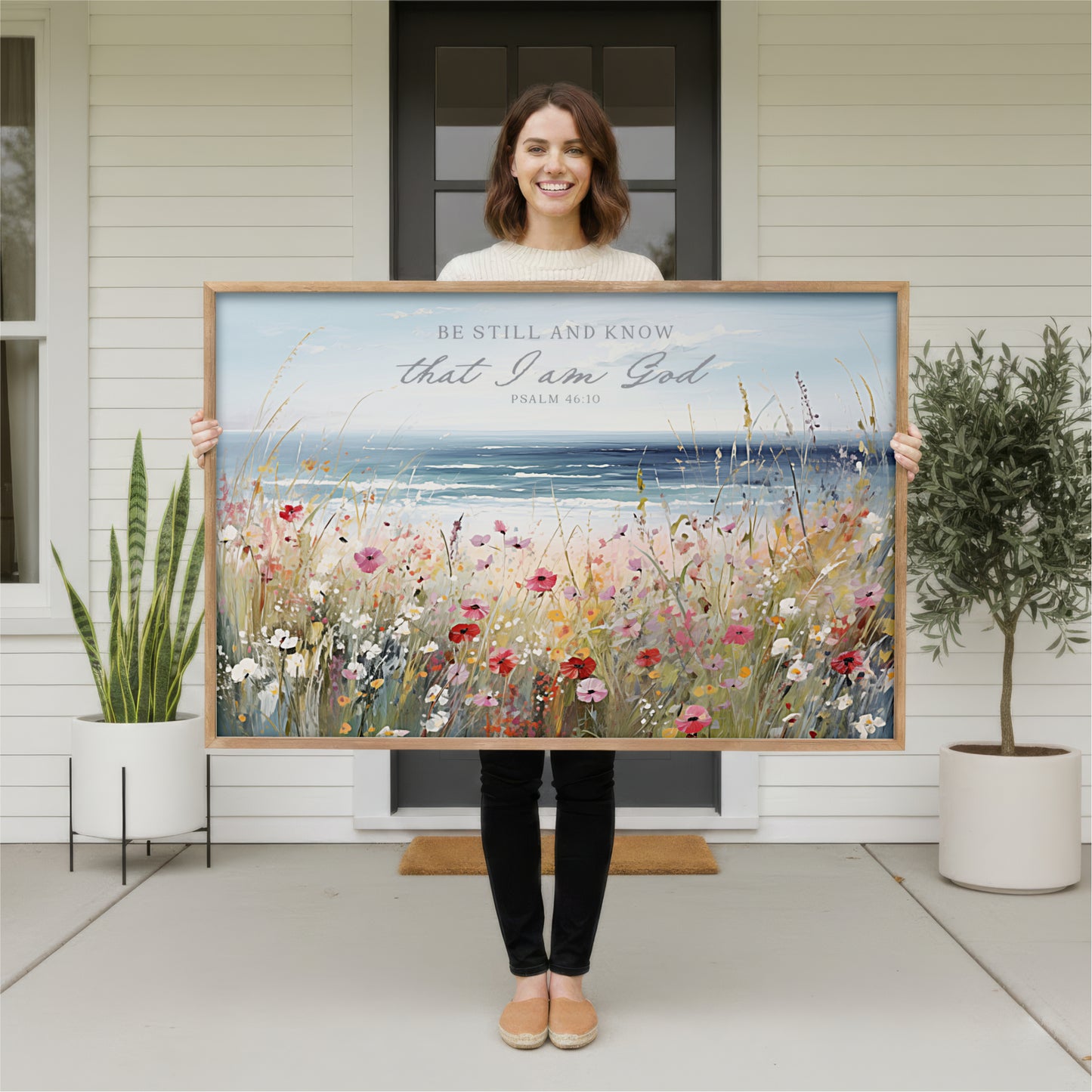 A woman stands in front of a white house, holding a large framed painting of a field of flowers.