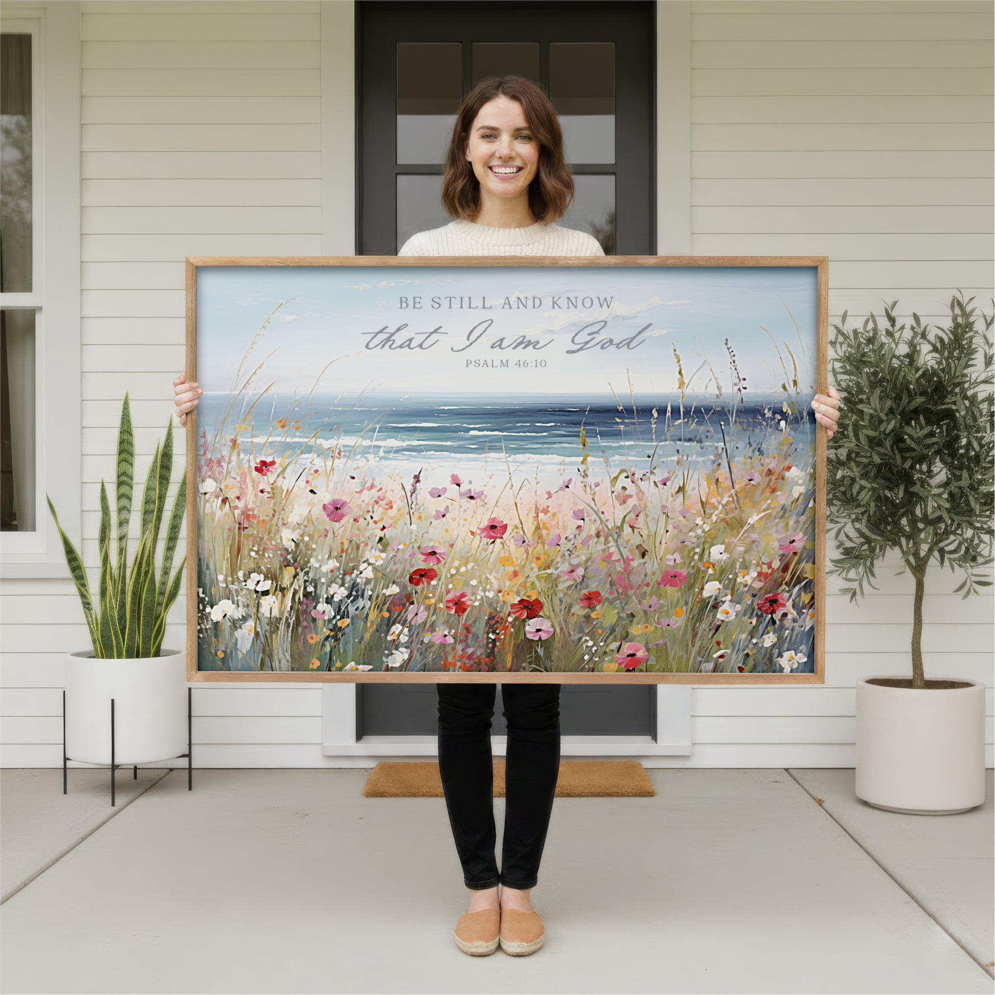 A woman stands in front of a white house, holding a large framed painting of a field of flowers.
