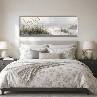 a serene bedroom scene with a large bed adorned with a floral comforter and pillows, flanked by two nightstands with lamps. Above the bed, a large framed picture of a beach landscape with tall grasses and a cloudy sky is displayed on the wall.