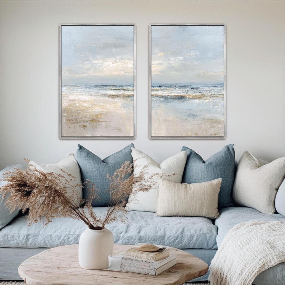 a cozy living room with a blue couch adorned with white and blue throw pillows, a wooden coffee table, and two framed paintings of a serene beach scene hanging on the wall above the couch.