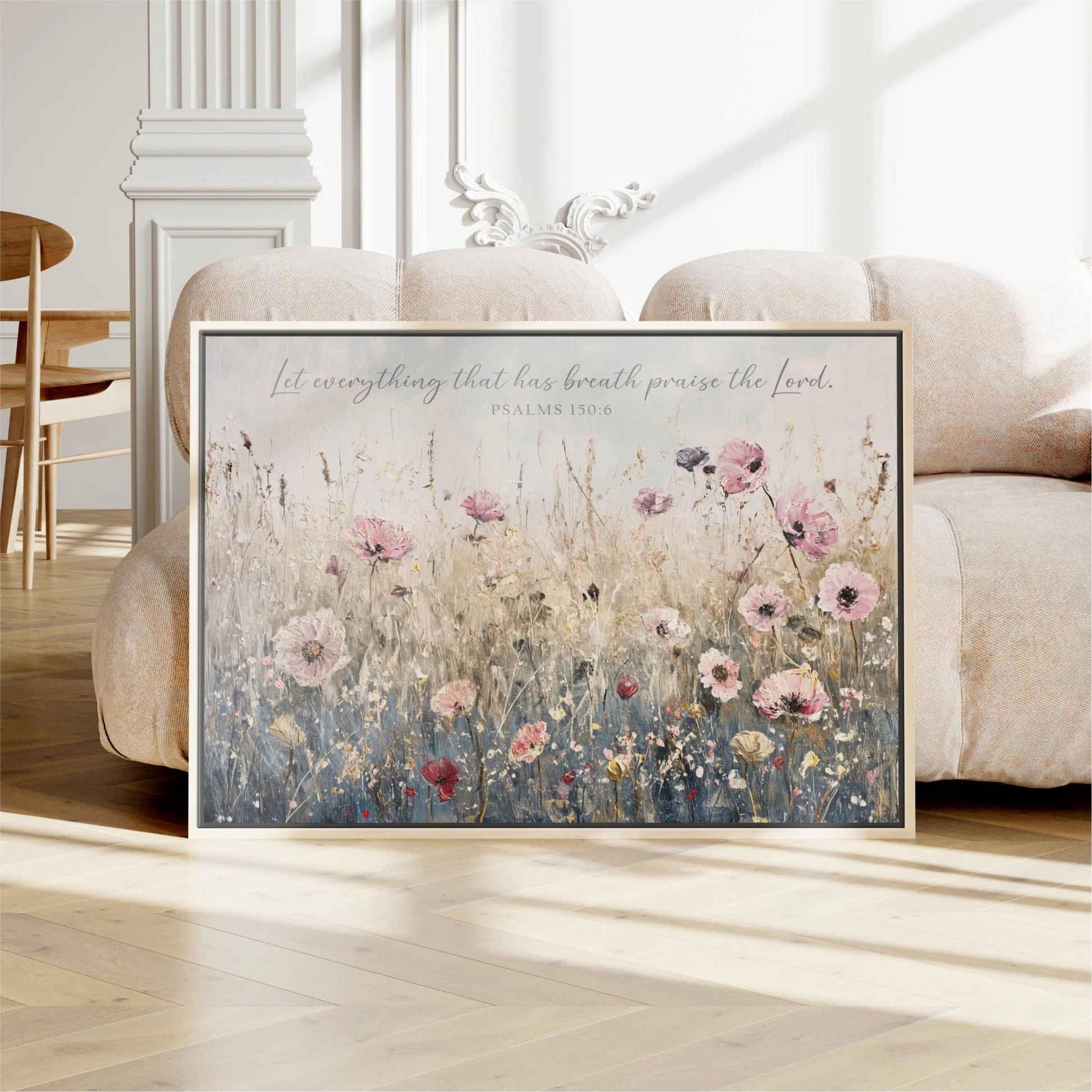 A framed painting of a field of flowers is displayed on a wooden floor in a room with a white sofa and a wooden chair.