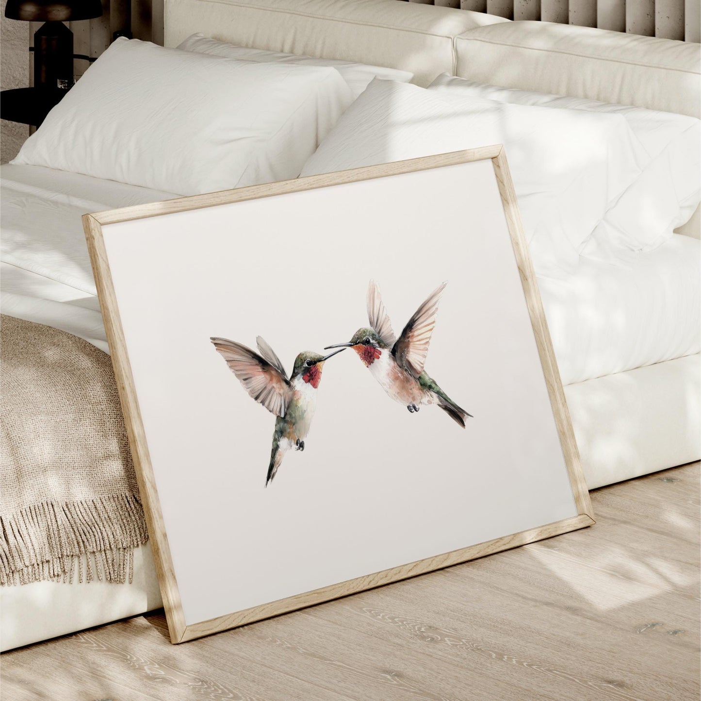 A framed print of two hummingbirds is displayed on a bed, with a white bedspread and pillows in the background.