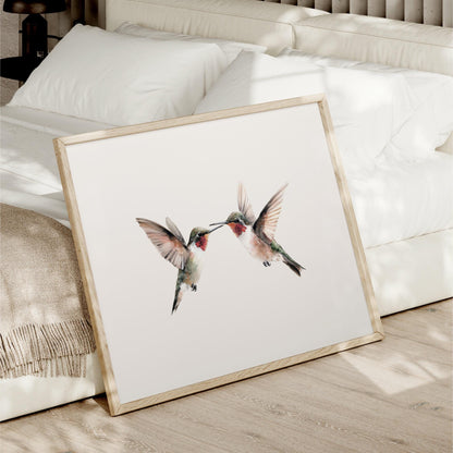 A framed print of two hummingbirds is displayed on a bed, with a white bedspread and pillows in the background.