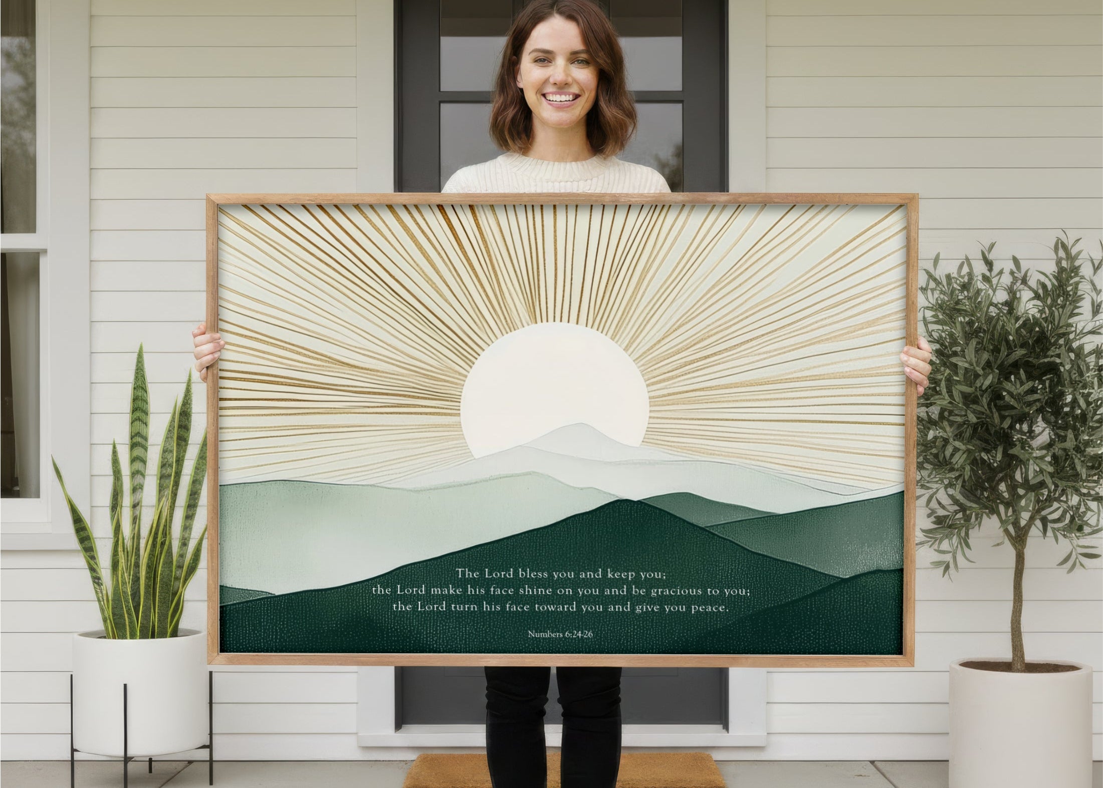 A woman stands in front of a white house, holding a large framed print of a landscape with a sun and mountains.