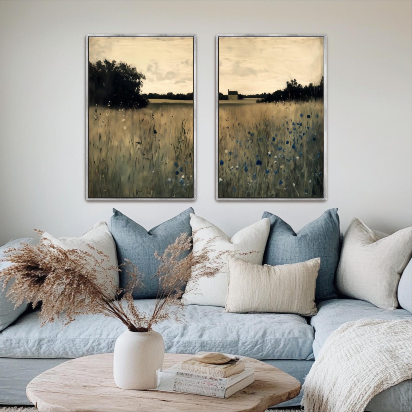 a cozy living room with a blue couch adorned with white and blue pillows, a white vase with dried flowers, and two framed landscape paintings hanging on the wall above the couch.