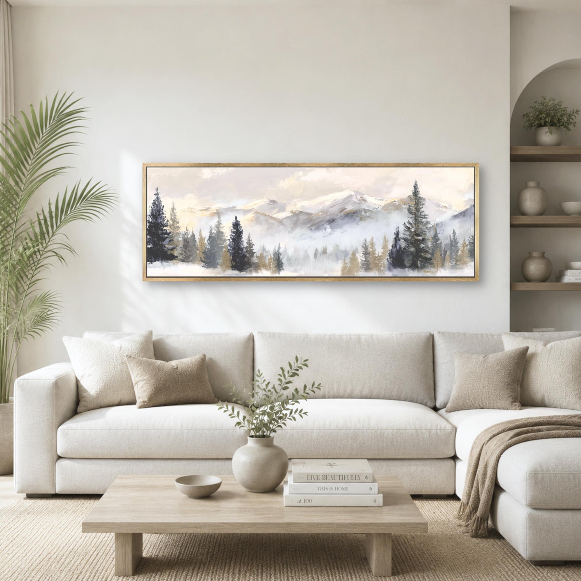 a living room with a large painting of a mountain landscape hanging on the wall above a white sofa. The sofa is positioned in front of a coffee table, which has a vase of greenery and a stack of books on it. The room also includes a potted plant and a bookshelf, creating a cozy and inviting atmosphere.