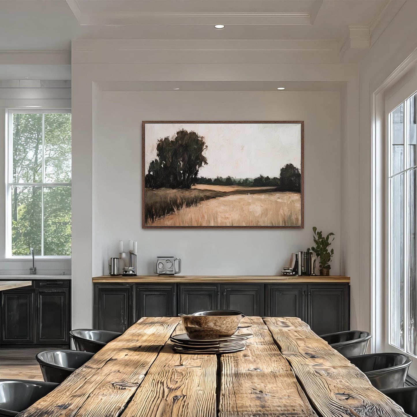 a dining room with a large wooden table surrounded by black chairs. Above the table, there is a framed painting depicting a rural landscape with trees and a field. The room has white walls and a window that allows natural light to enter the space.