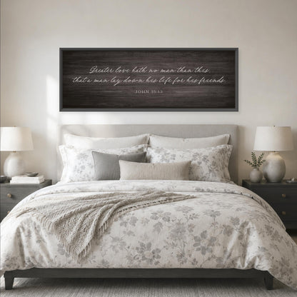 a bedroom with a large bed, a wooden headboard, and a framed quote above it. The bed is covered in a white and gray floral comforter, and there are two lamps on either side of the bed. The quote on the wall reads, "Greater love hath no man than this that a man lays down his life for his friends."